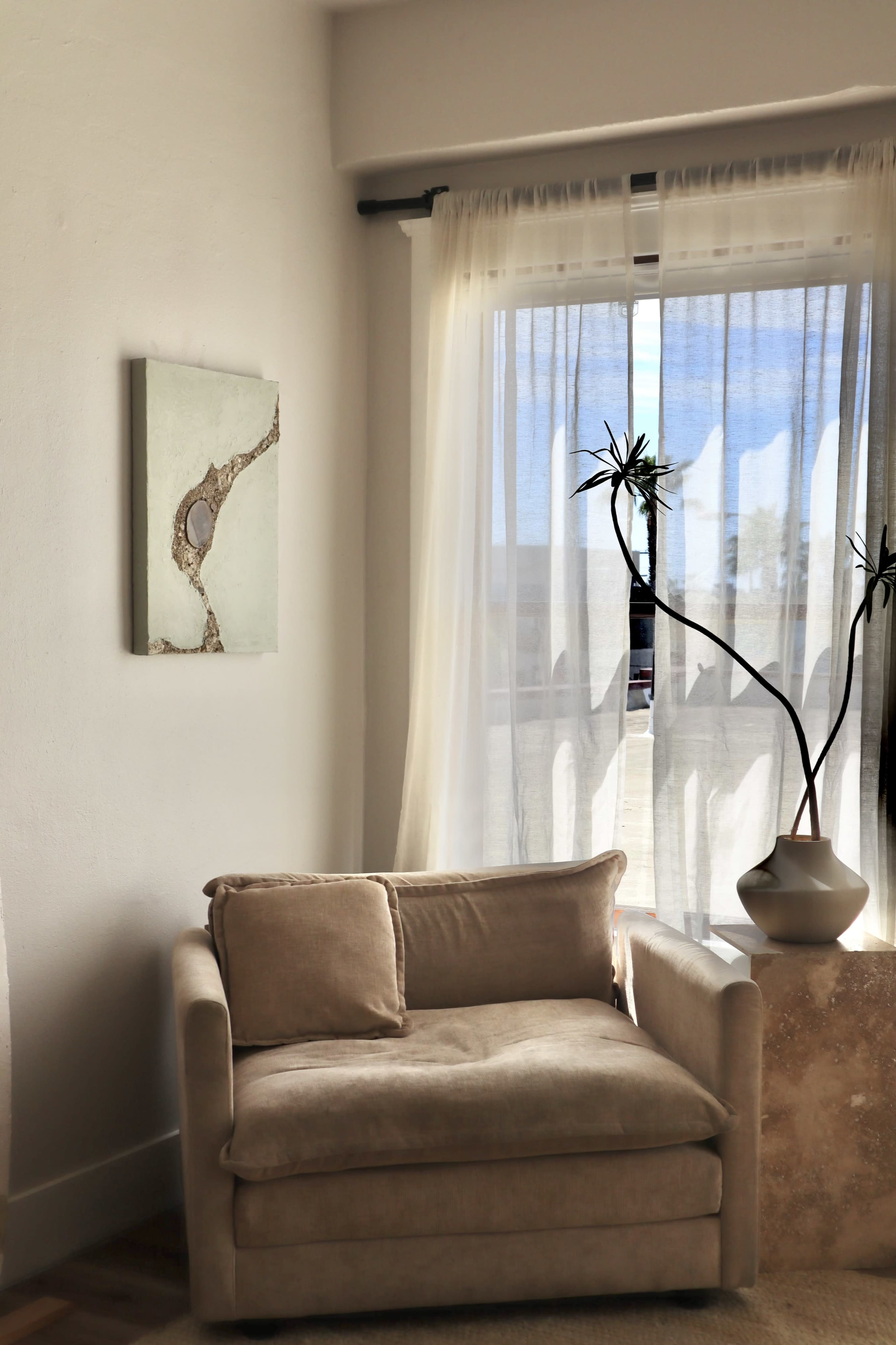 A beige armchair is positioned beside a window with sheer curtains, featuring a small potted plant on a stone side table.