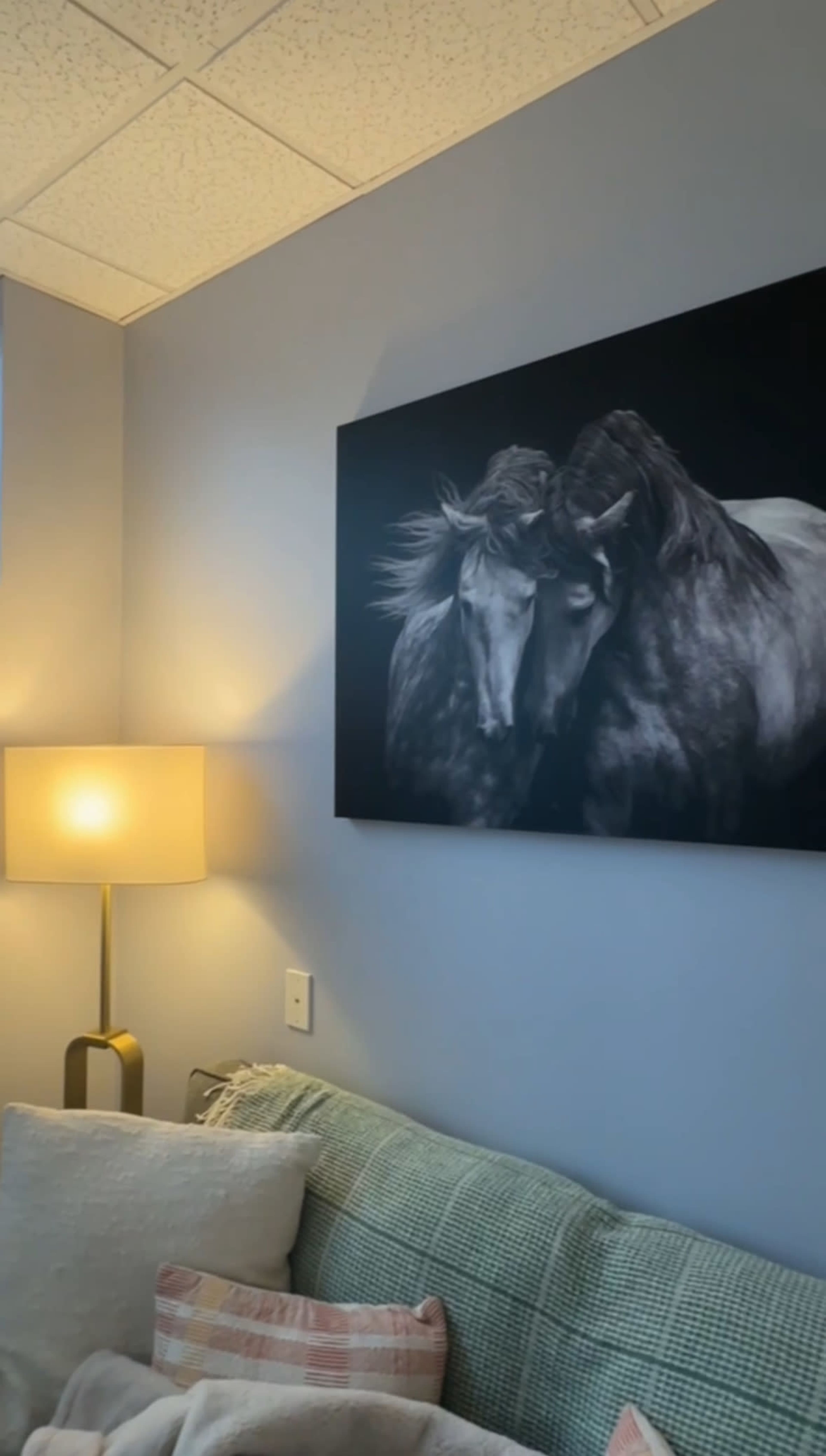 The image shows a cozy corner with a light-colored sofa, a lamp with a soft glow, and a large black-and-white painting of two horses on the wall.