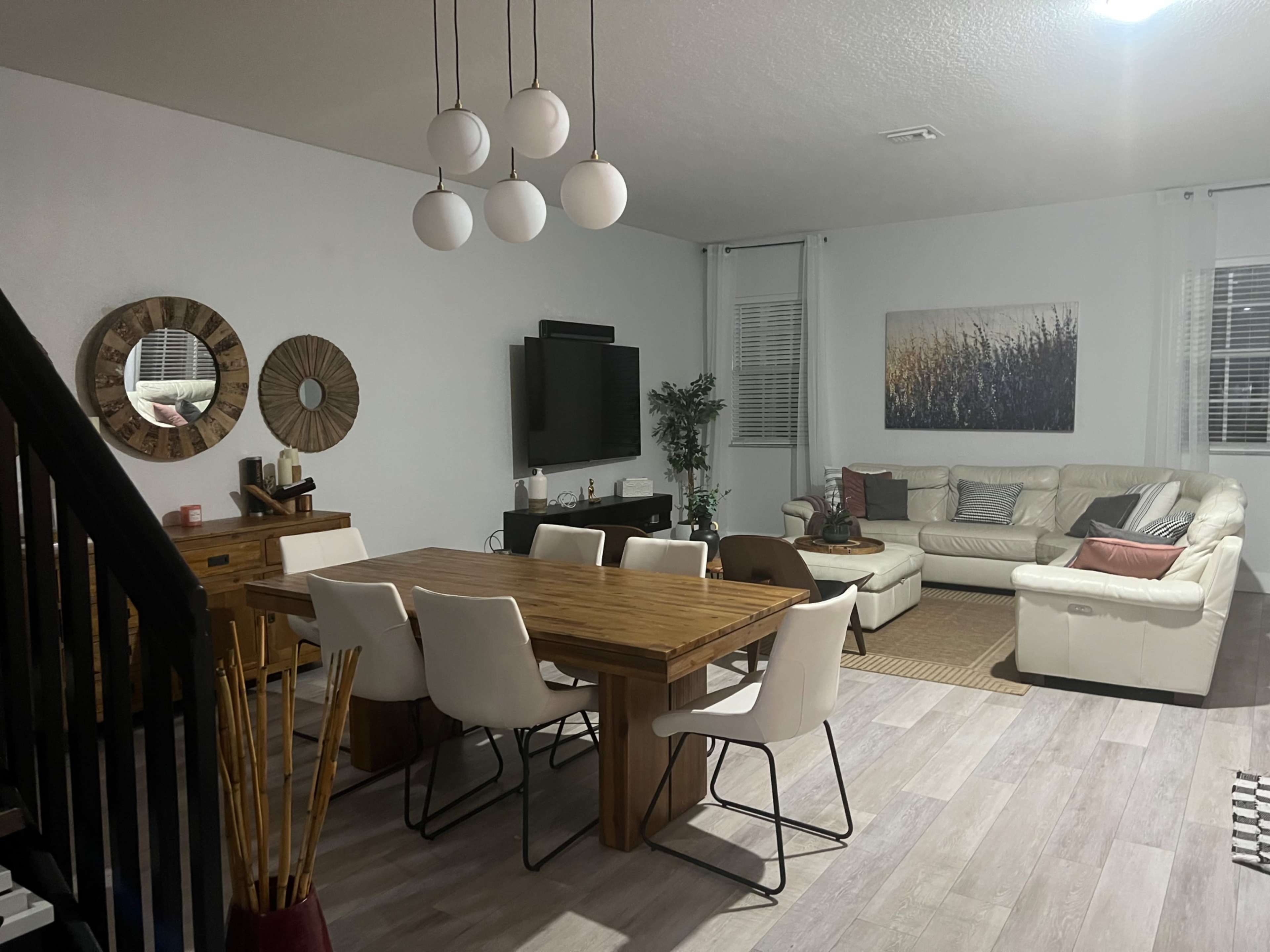 A modern living and dining area featuring a wooden dining table with white chairs, a cream-colored sectional sofa, and wall decorations including mirrors and artwork.
