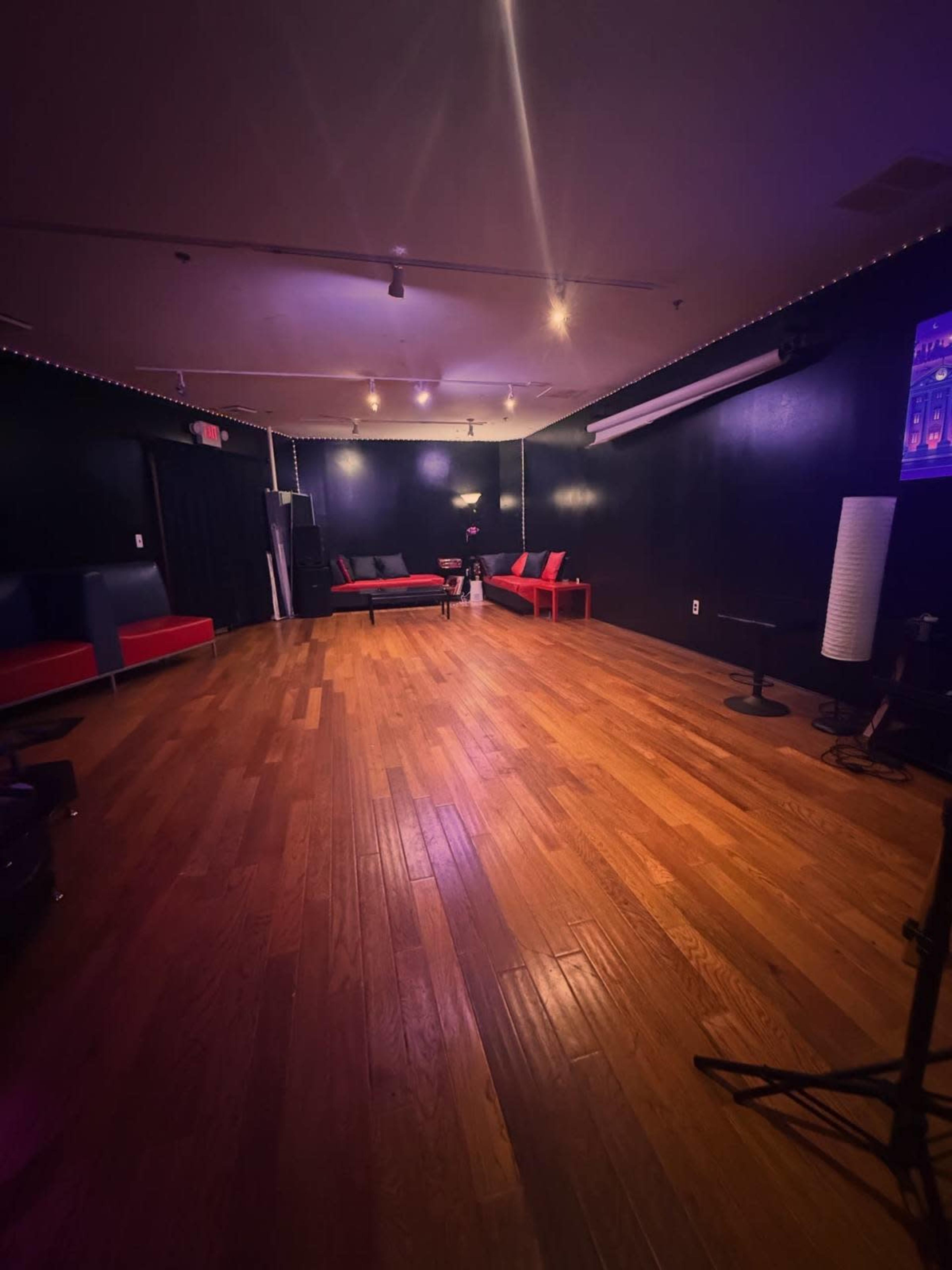 The image shows a dimly lit room with hardwood flooring and red and black furniture, including sofas and chairs arranged around the space.