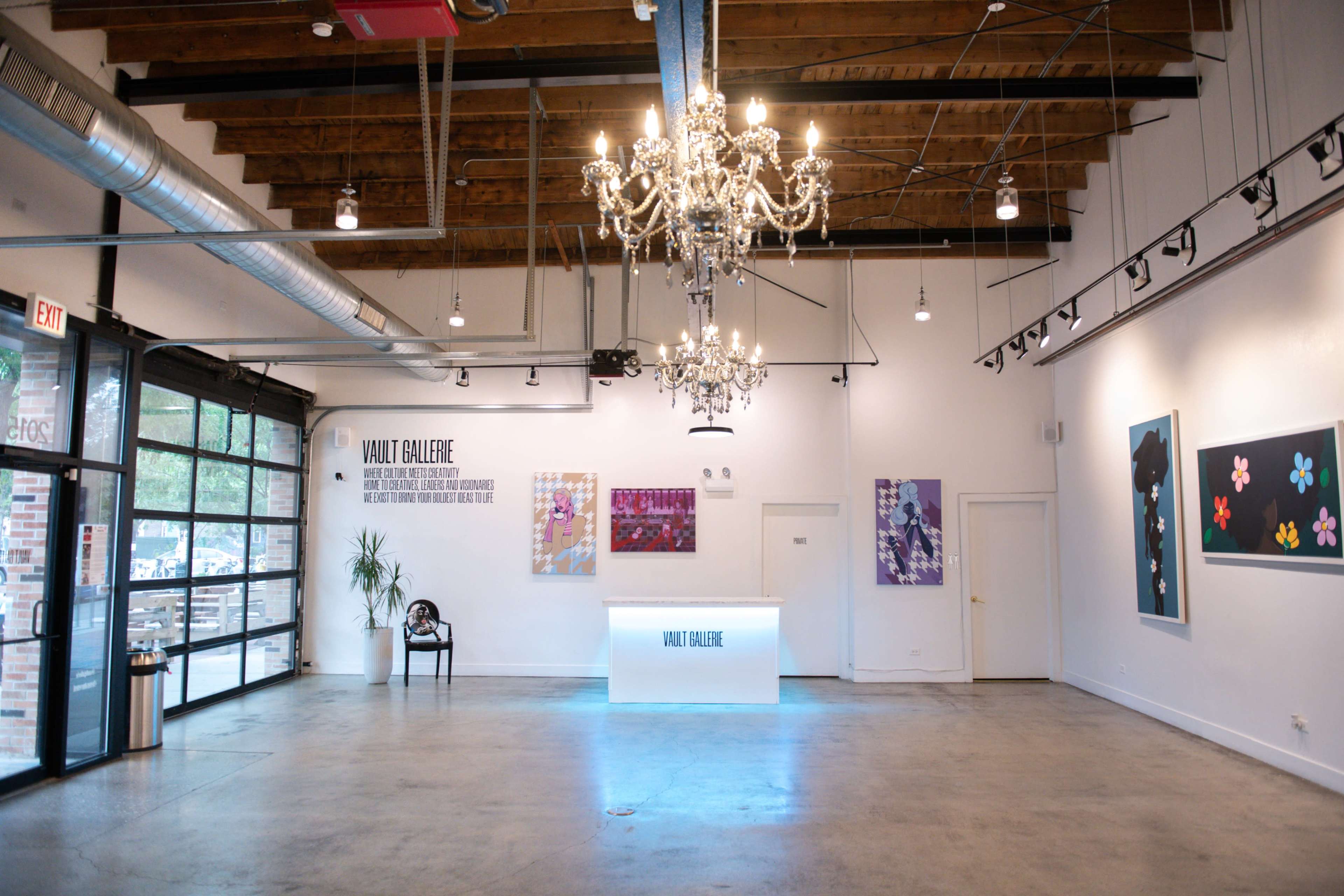 The image shows a modern gallery interior with a reception desk, a chandelier, and colorful artwork displayed on the walls.