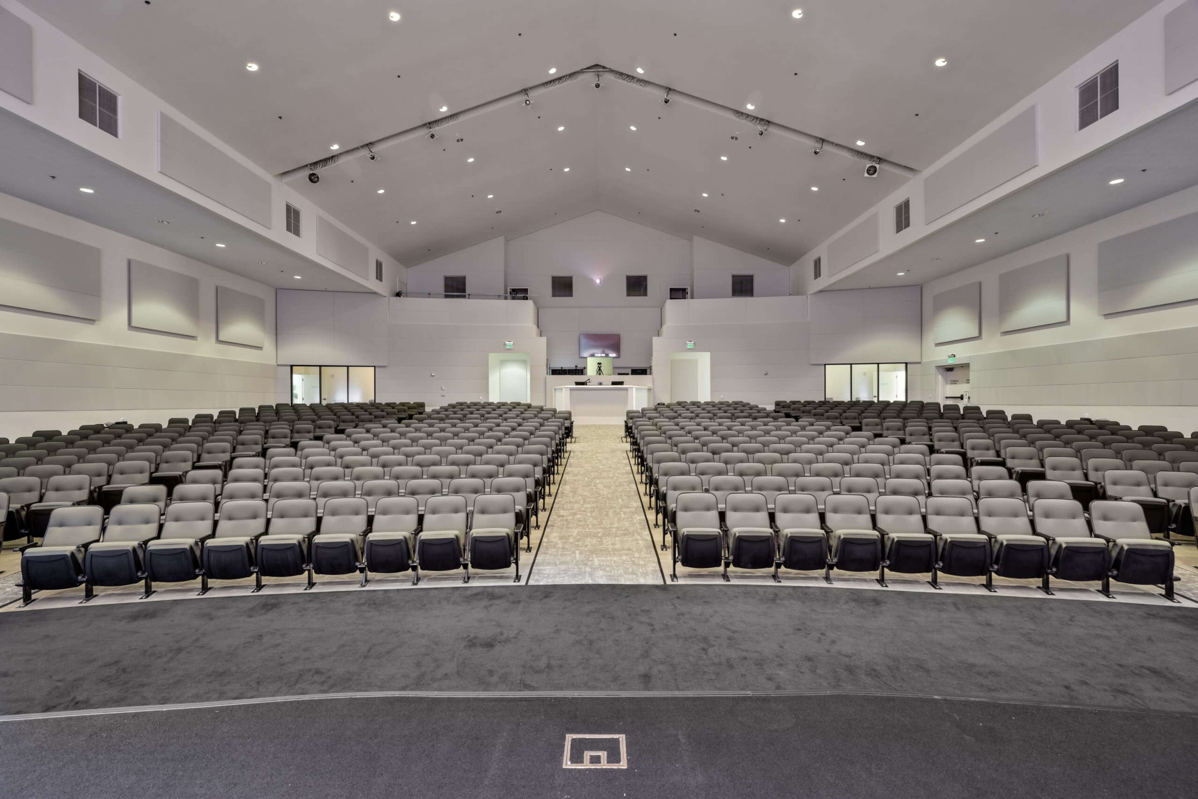 The image shows a spacious, empty auditorium with rows of gray chairs arranged facing a stage at the front.
