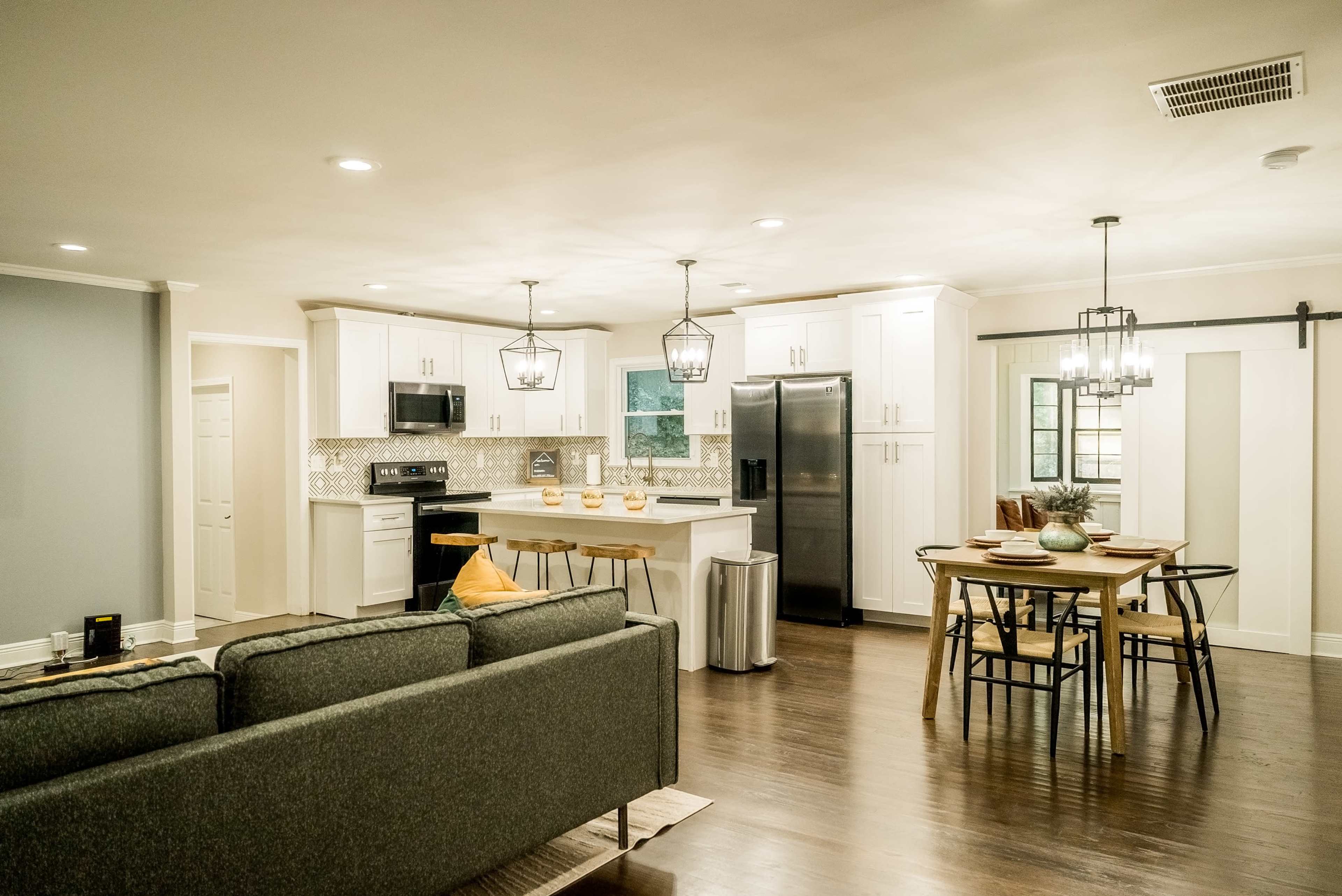 A modern kitchen with white cabinets and black appliances opens into a dining area featuring a table surrounded by chairs and a sofa in the foreground.