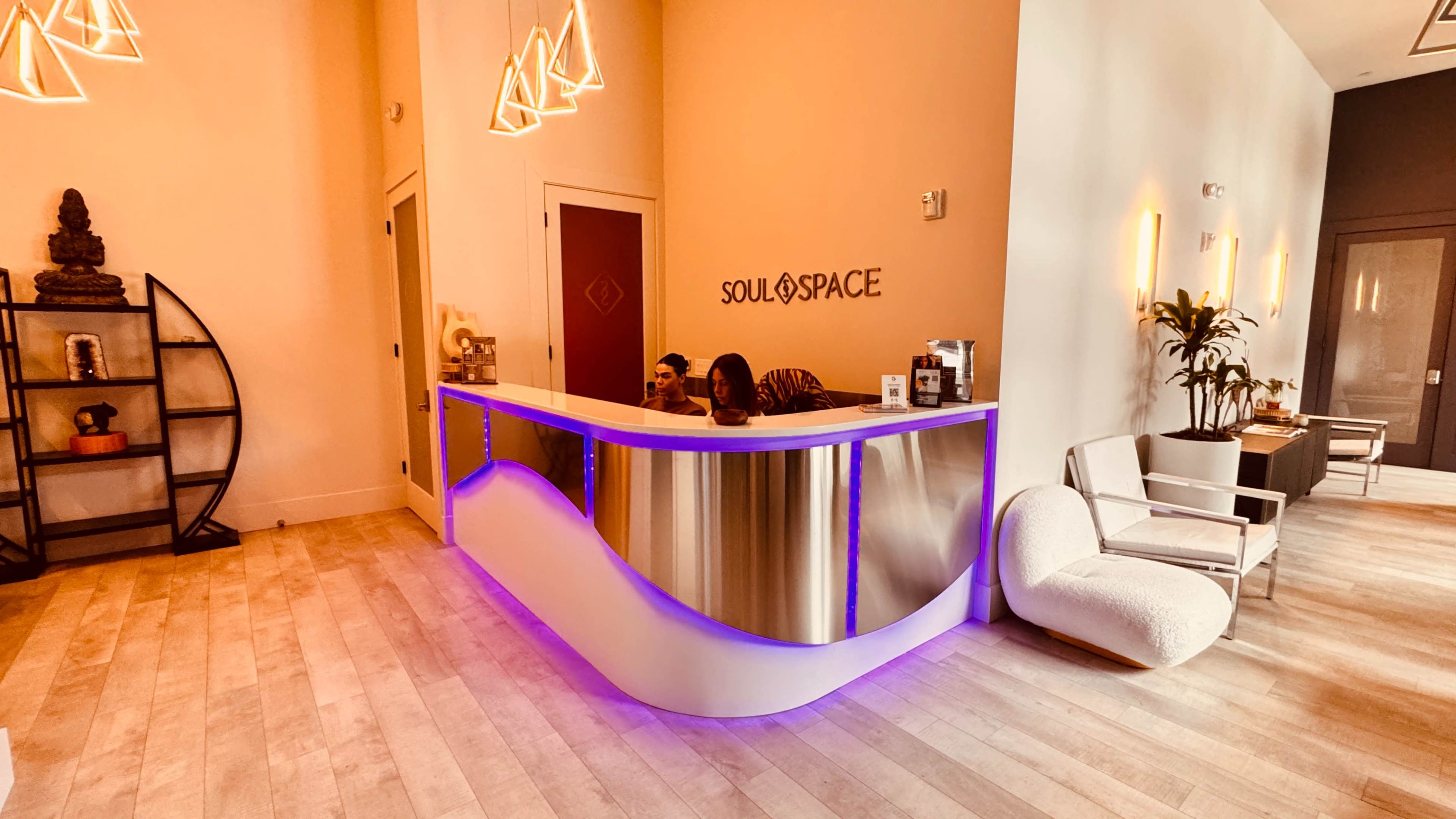 The image shows a modern reception area with a curved front desk illuminated by purple lighting, featuring two staff members behind the counter.