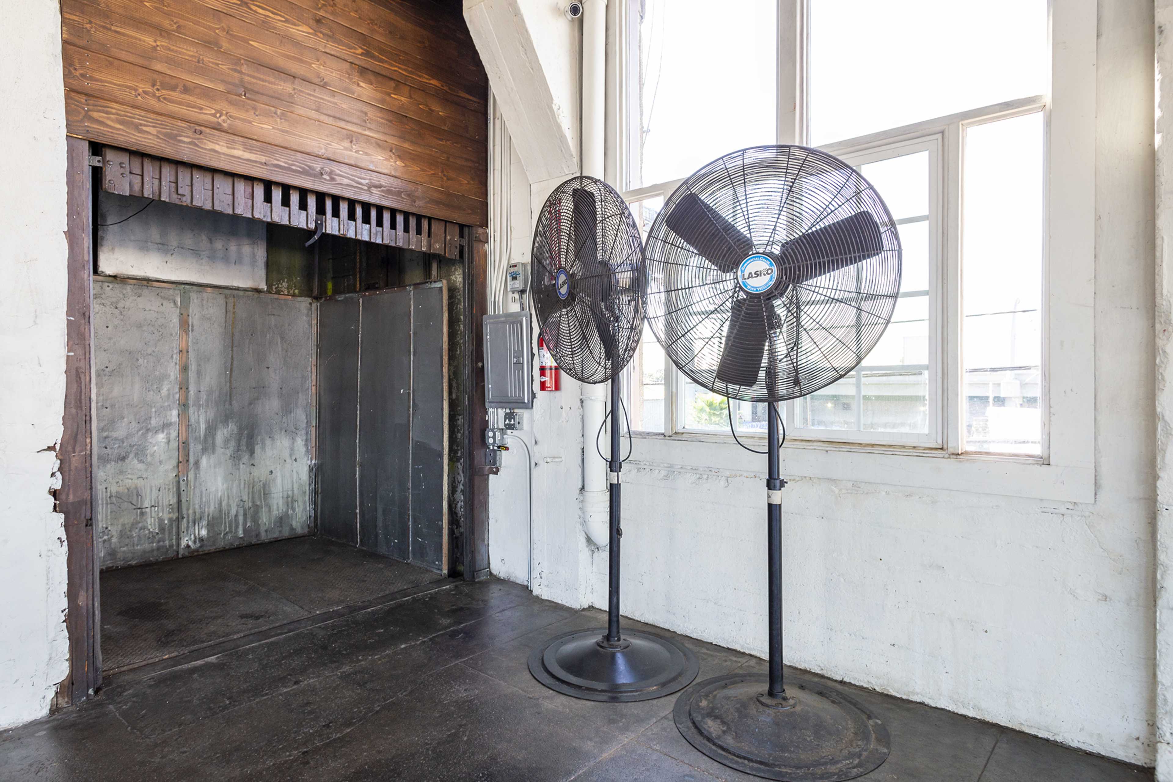 Two large industrial fans stand next to an open elevator door in a bright, airy room with large windows.