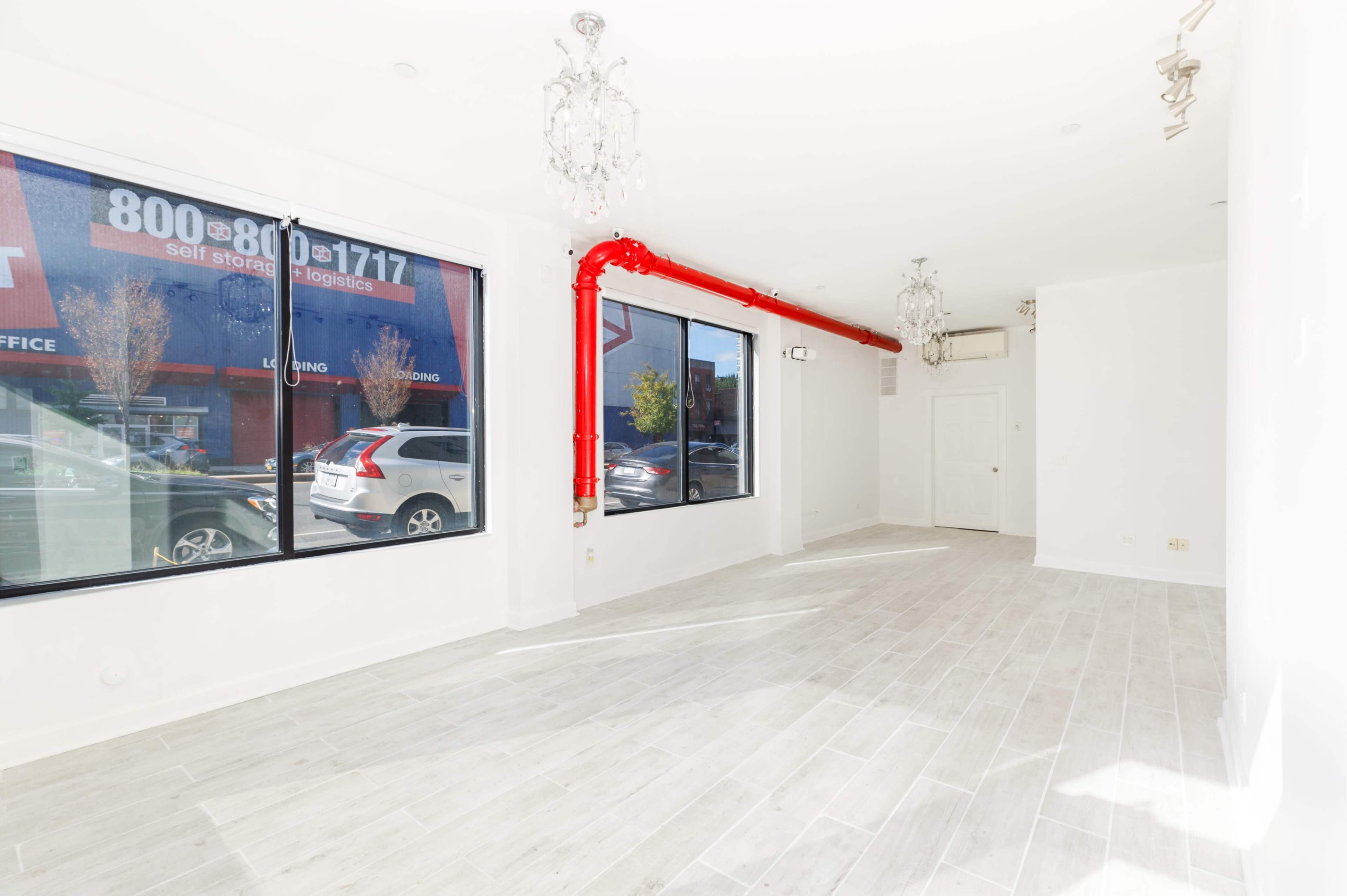 An empty retail space with white walls, light-colored flooring, and a prominent red pipe along the ceiling.