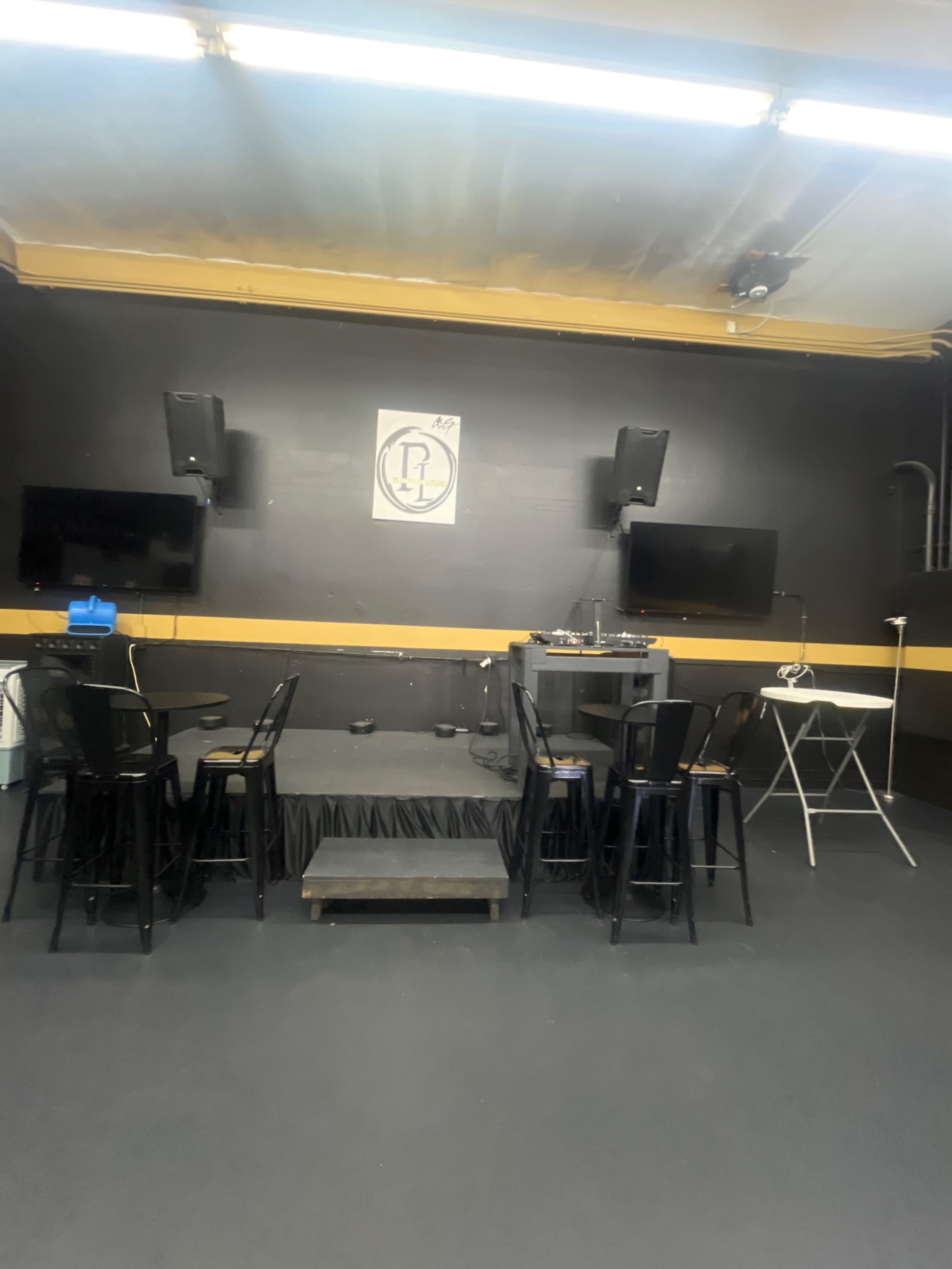 The image shows a small stage area with a black backdrop, two speakers mounted on the walls, two television screens, and several black chairs arranged around a power outlet.