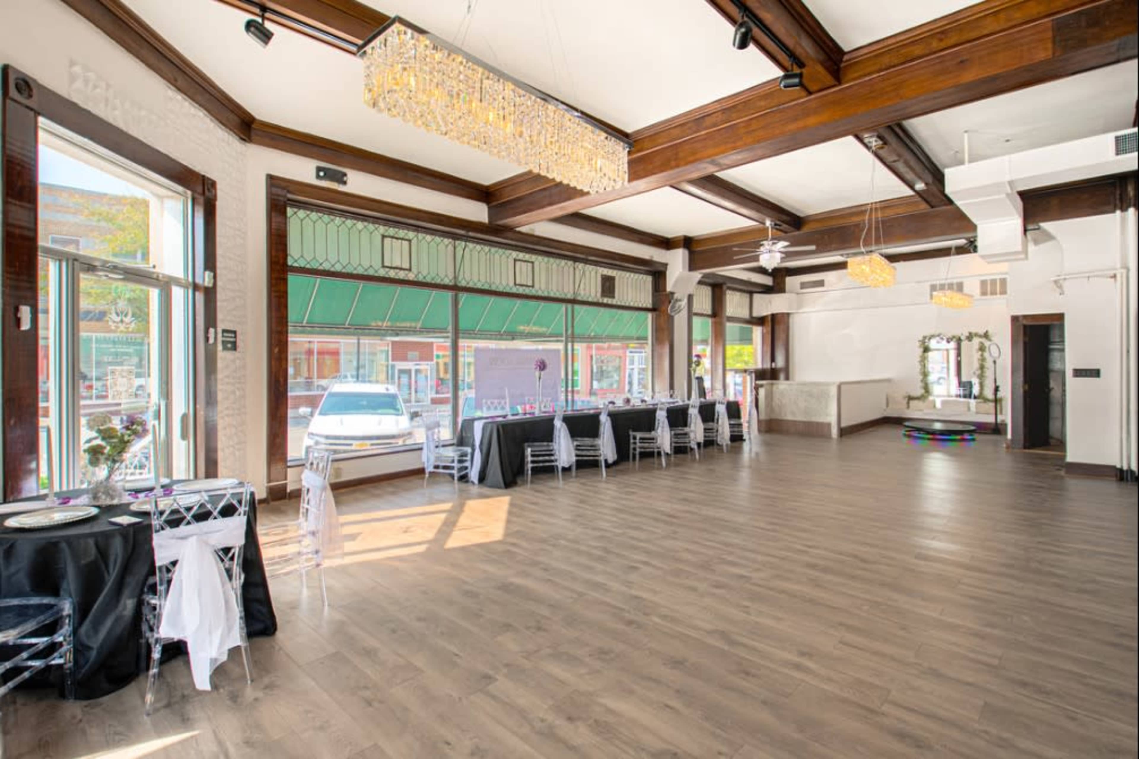 The interior of a spacious venue features hardwood floors, large windows, and a combination of black and white table settings.