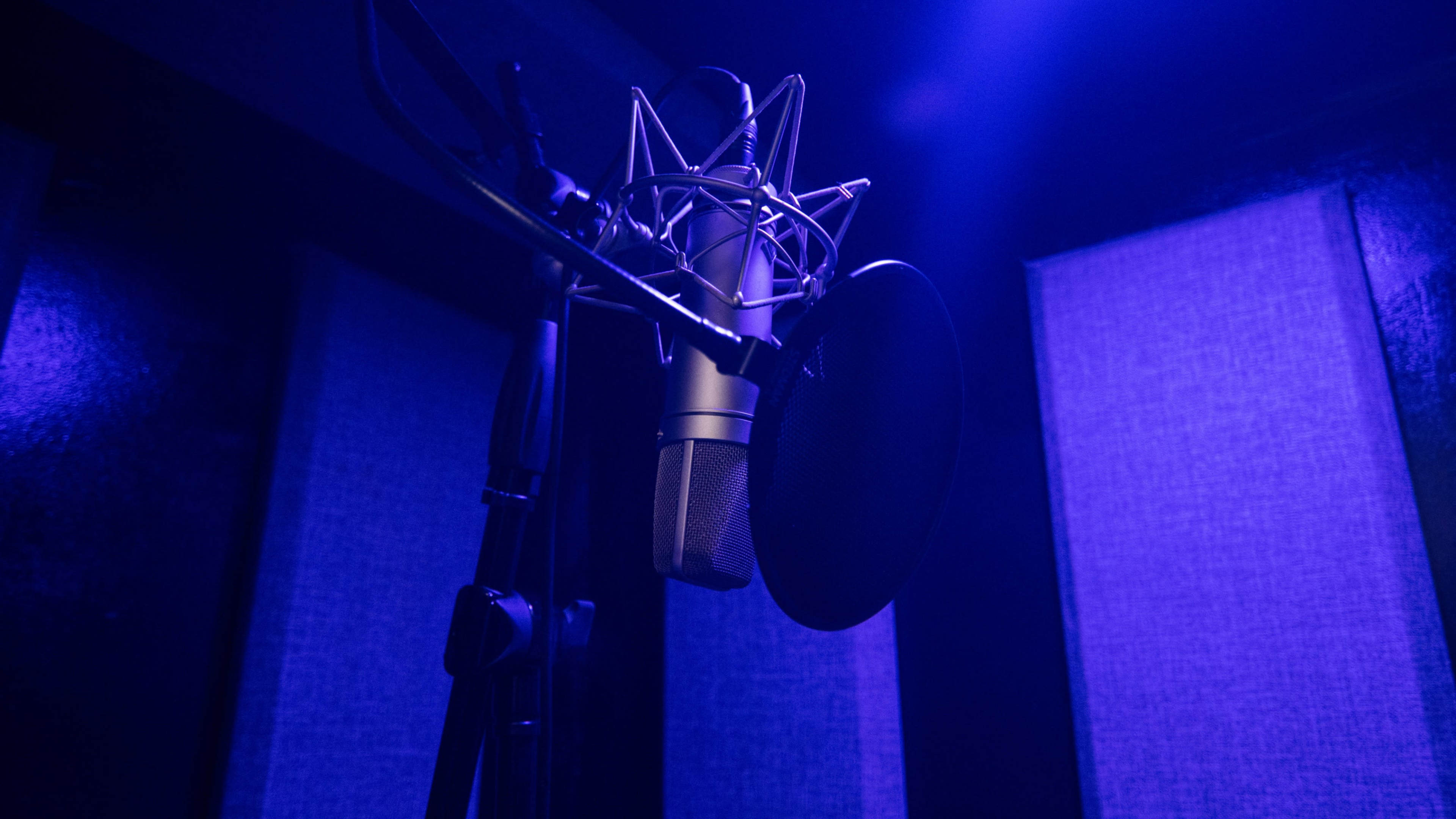 A microphone stands on a stand inside a soundproof recording studio with purple lighting.