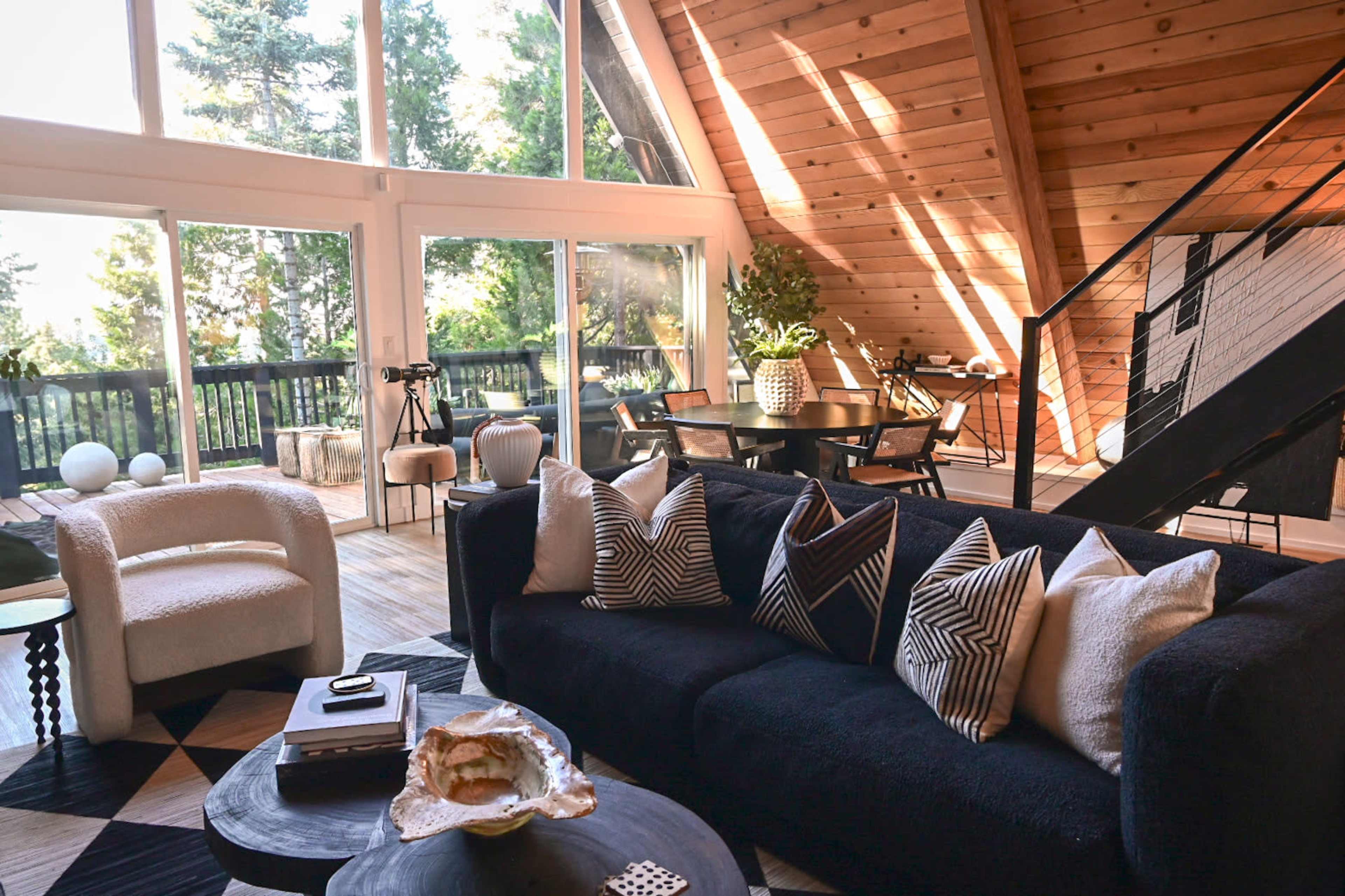 A modern living room with a black sofa adorned with patterned pillows, a round table, and large windows that offer a view of trees outside.