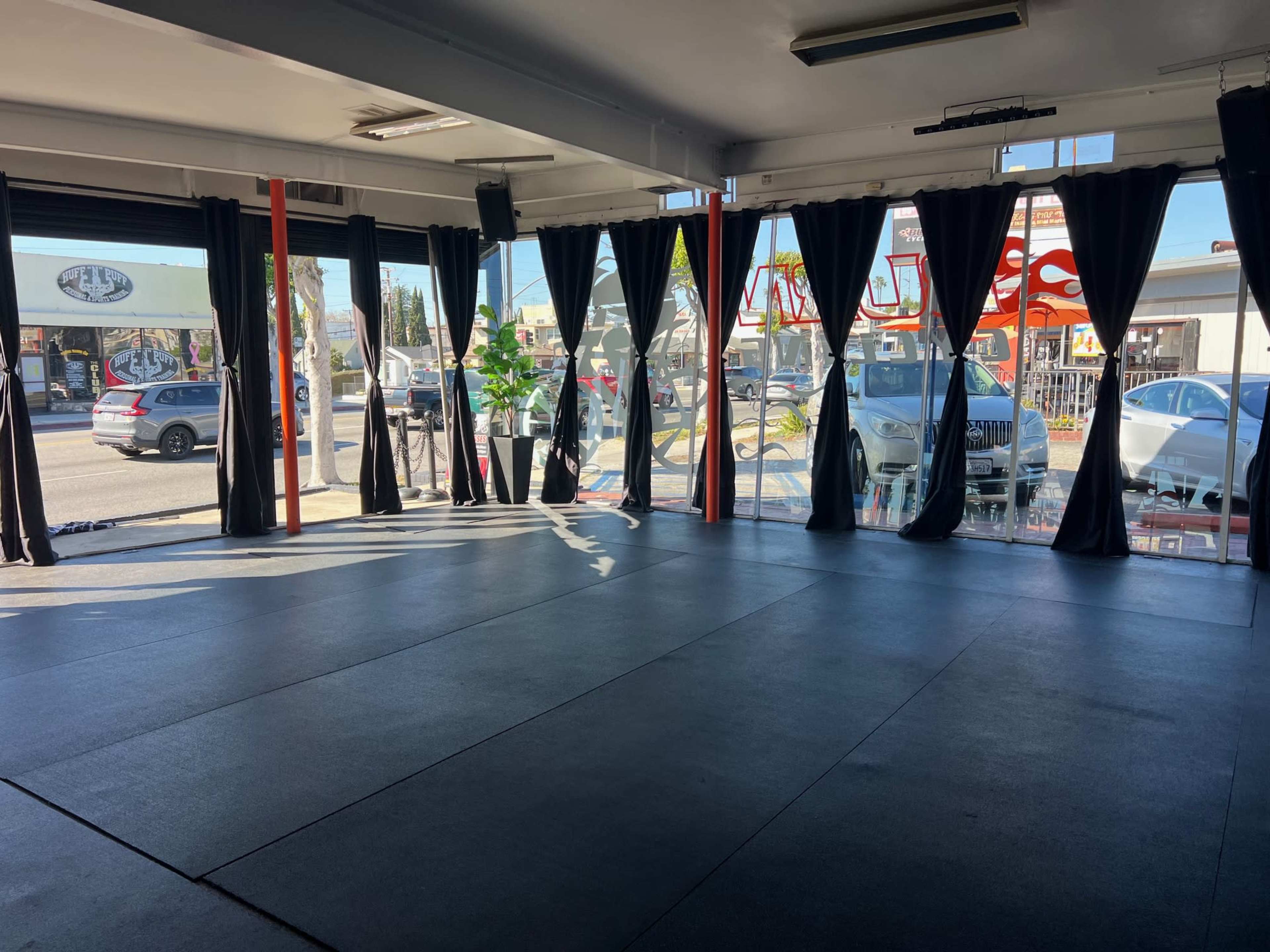 The image shows an empty gym or exercise space with black mats on the floor and large windows covered by dark curtains, allowing a view of vehicles outside.