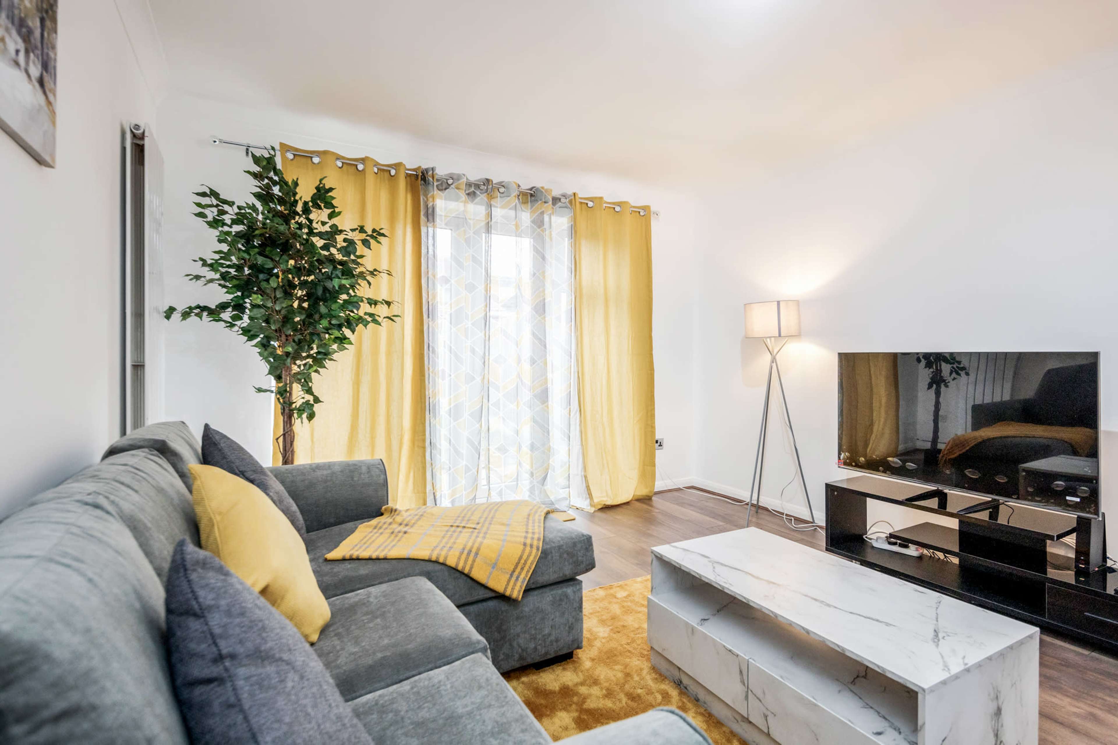 A living room with a gray sectional sofa, a marble coffee table, yellow curtains, a floor lamp, and a large flat-screen TV.