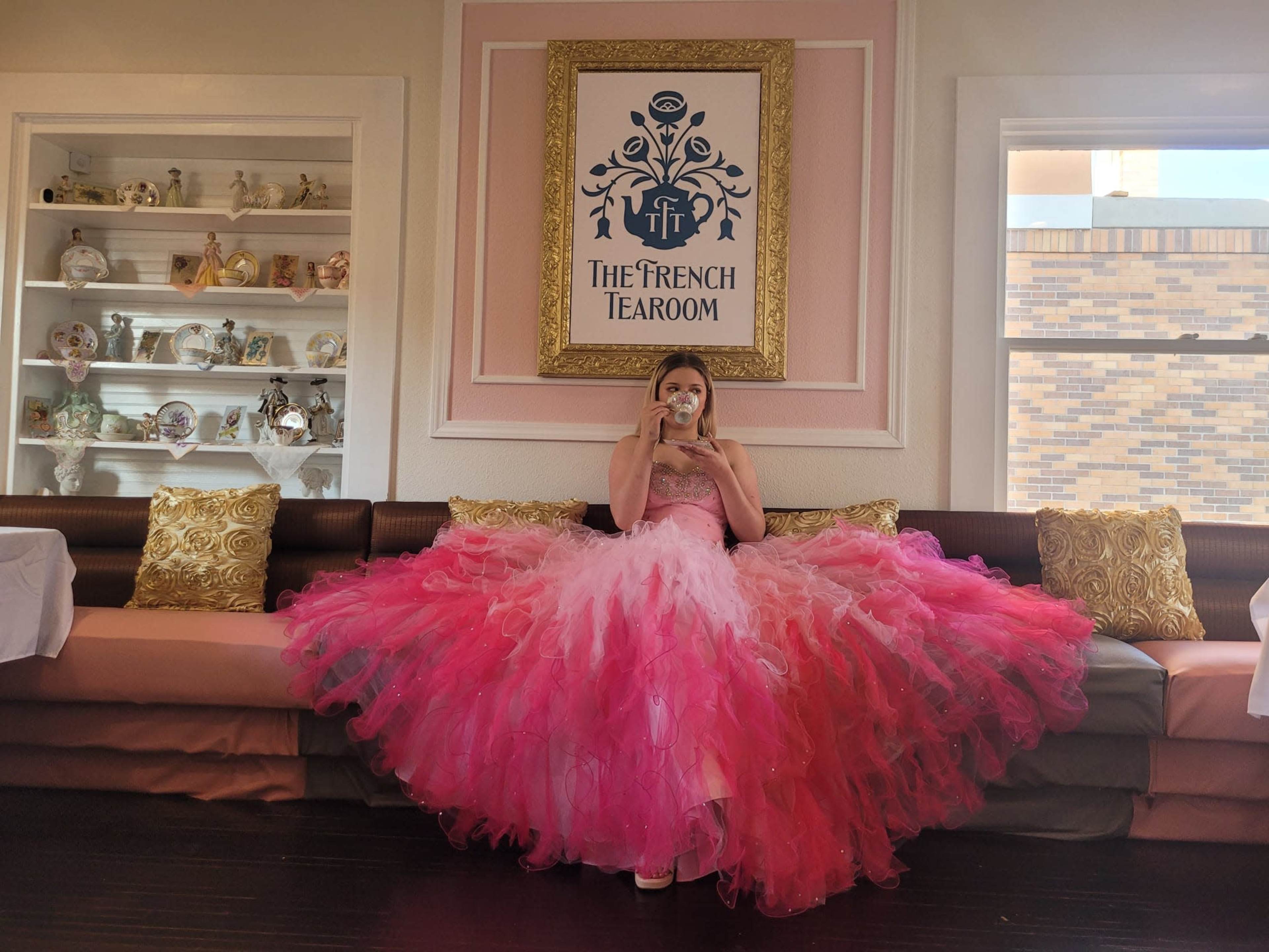 A person in a large pink and white dress sits at a table in a tea room decorated with shelves of decorative items.
