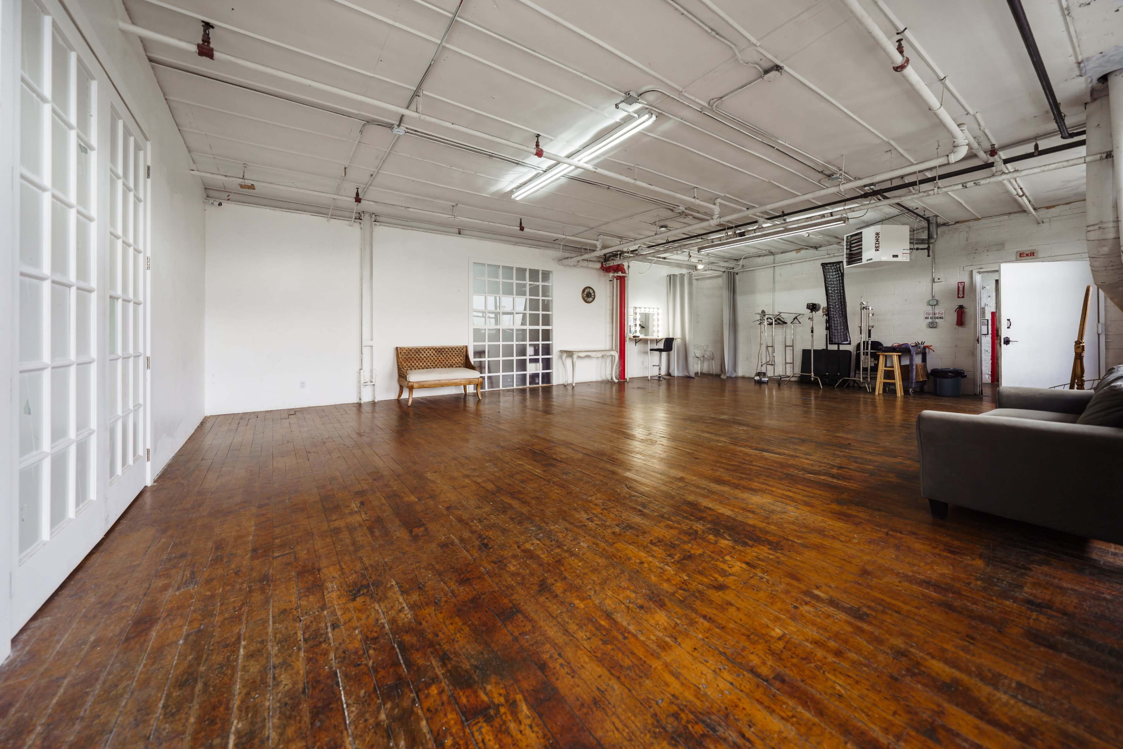A spacious, empty room with a wooden floor, large windows, and minimal furniture, including a couch and a bench.