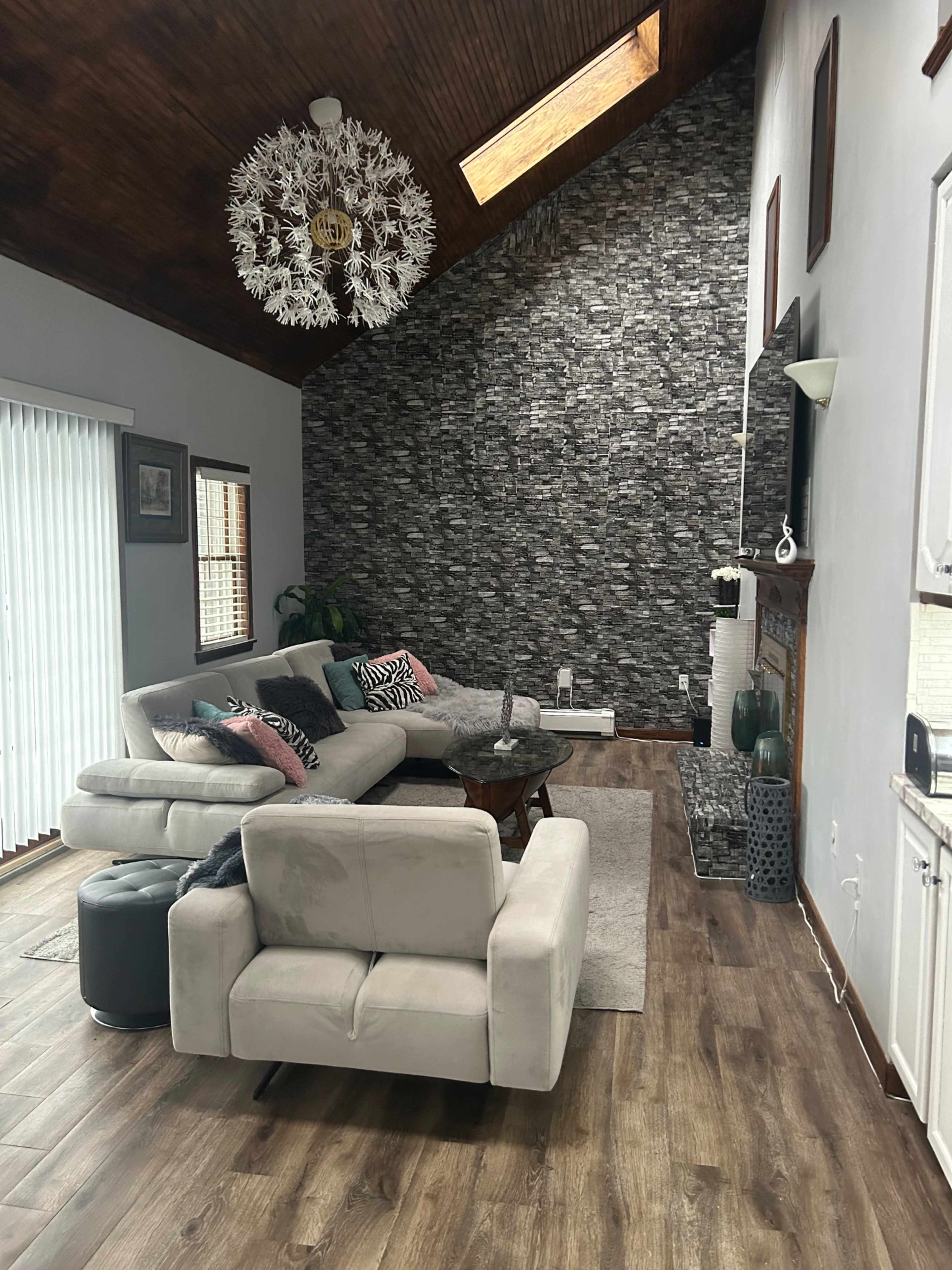 The image shows a stylish living room featuring a gray sectional sofa with colorful cushions, a circular coffee table, and a stone accent wall, all under a wooden-beamed ceiling with a skylight.