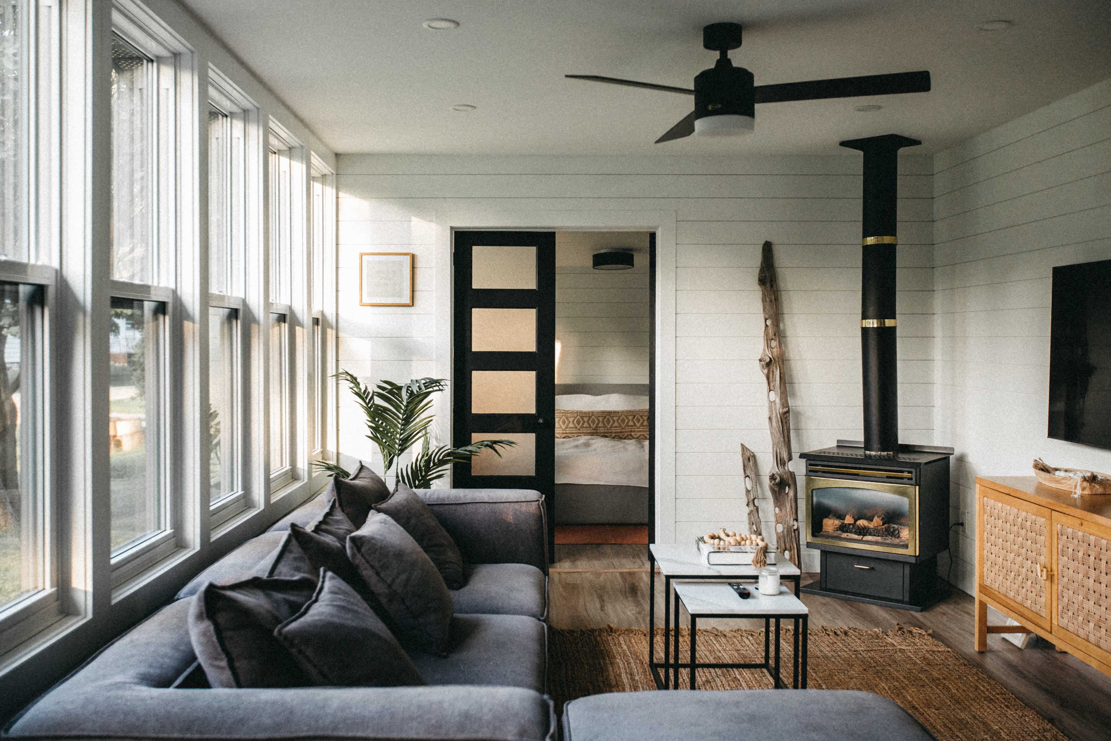 A modern living room features large windows, a gray sofa with decorative pillows, a black ceiling fan, and a wood stove, with a doorway leading to a bedroom in the background.