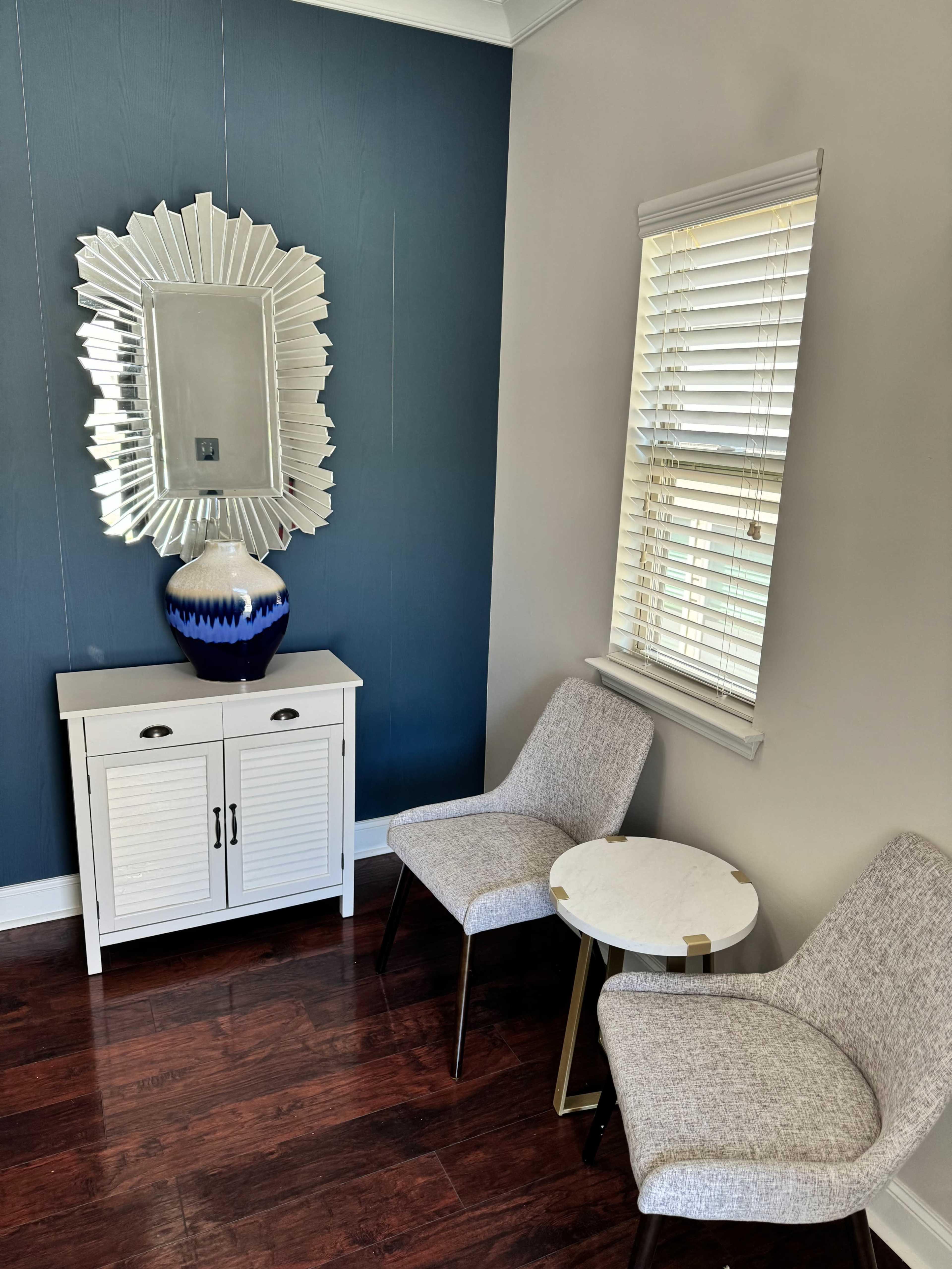 The image shows a small corner with two chairs, a round table, a cabinet, a decorative mirror, and a vase on a surface, set against a blue wall and hardwood flooring.