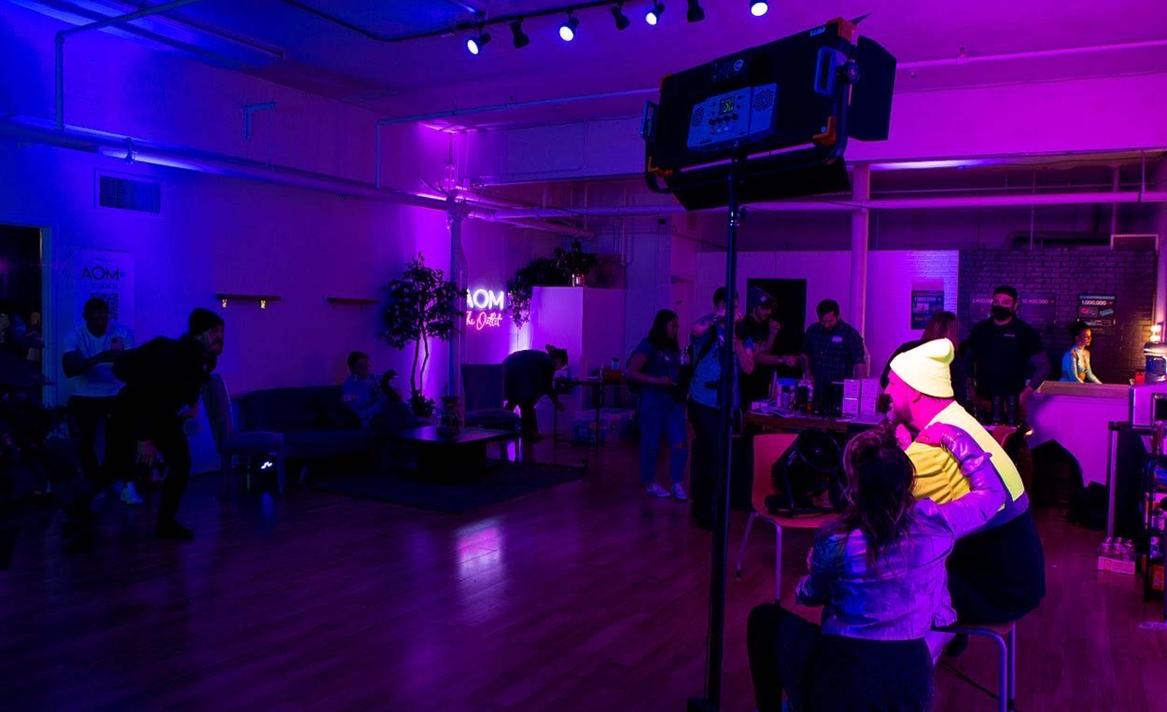 A group of people is gathered in a colorful room illuminated by purple and pink lights, with a mix of seating areas, as one person styles another's hair.