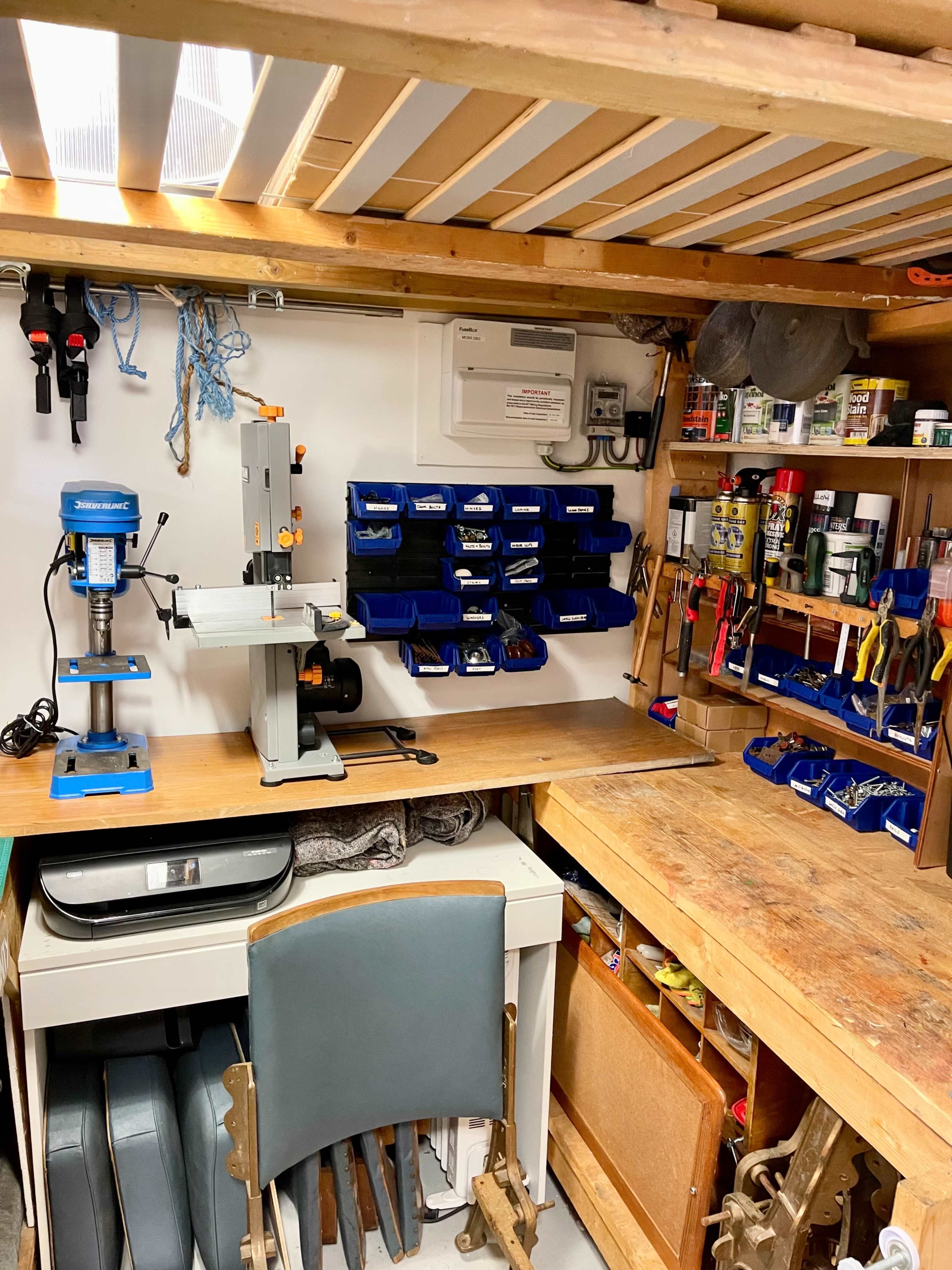 A workshop with various tools and equipment organized on shelves, including a scroll saw, a drill press, and storage bins for small items.
