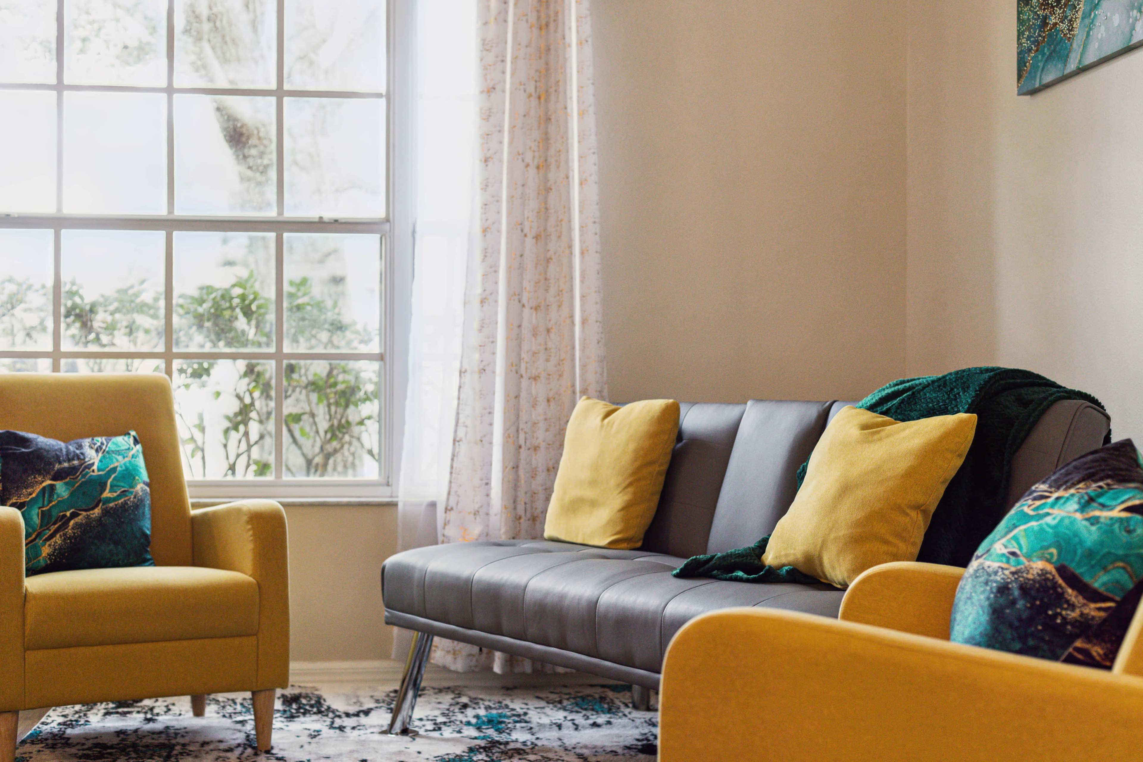 A cozy living room features a gray sofa and two yellow armchairs, with decorative pillows and a large window allowing natural light to enter.