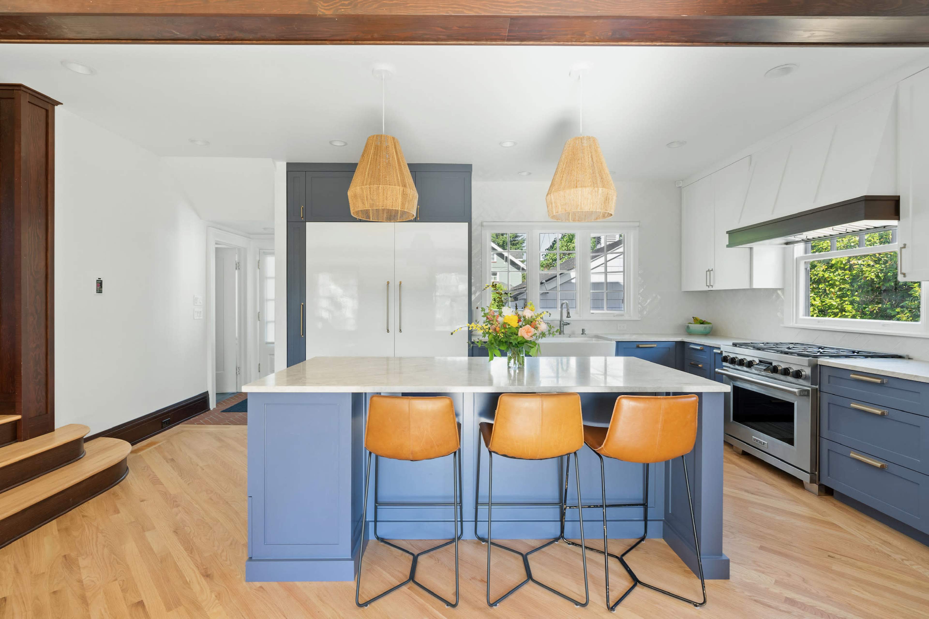 A modern kitchen features a blue island with three leather bar stools, pendant lights above, and large windows allowing natural light to illuminate the space.