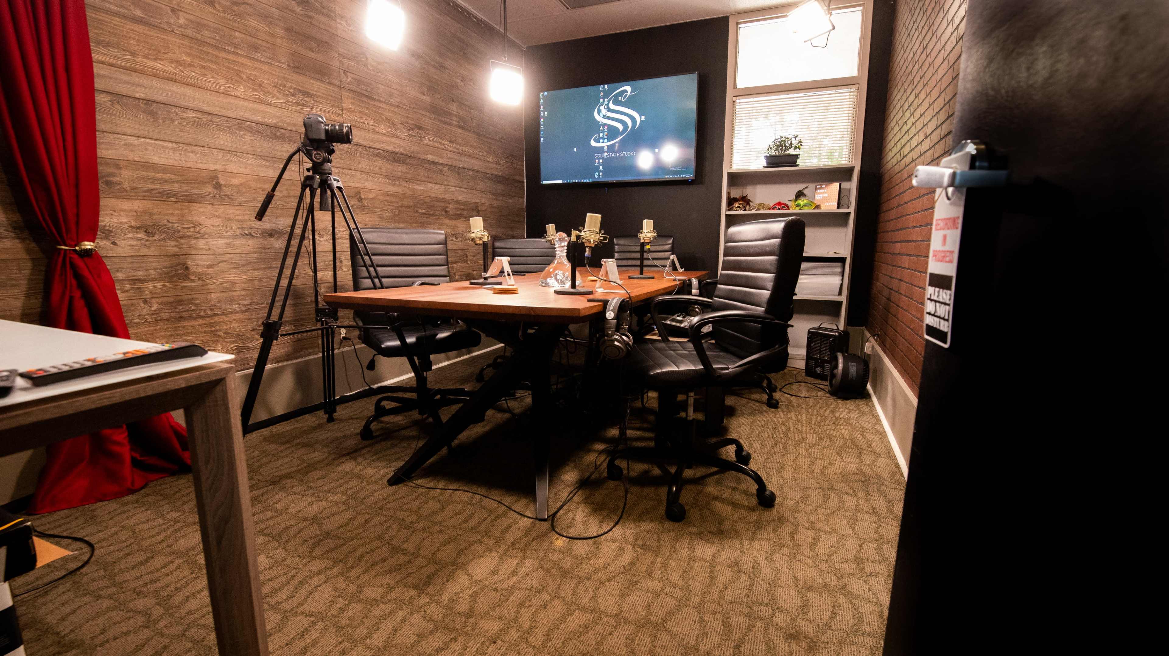 The image shows a small studio setting with a wooden table surrounded by black office chairs, a camera on a tripod, and a monitor displaying a logo on one wall.