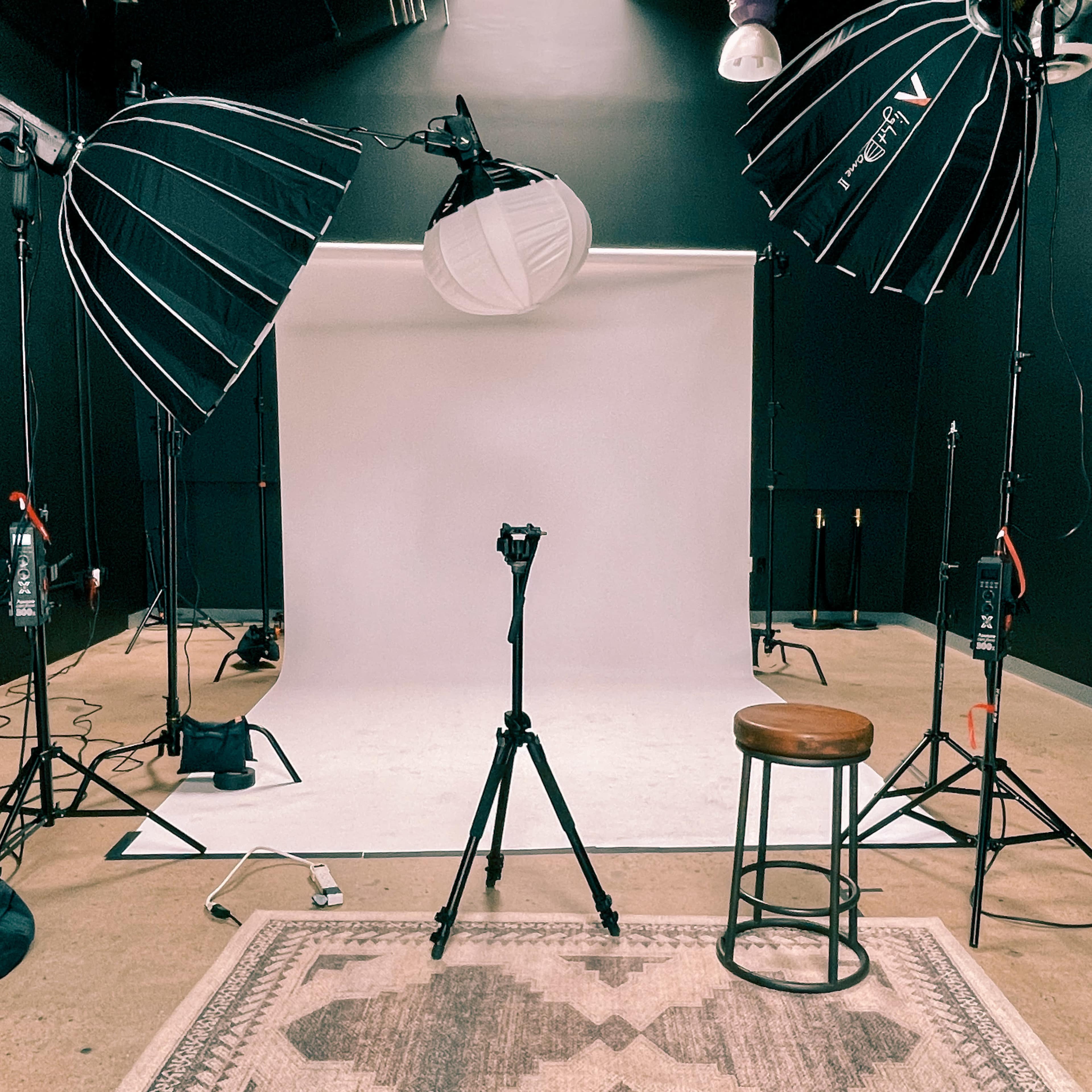 A photography studio setup with two large softbox lights, a white backdrop, a tripod, and a wooden stool on a patterned rug.