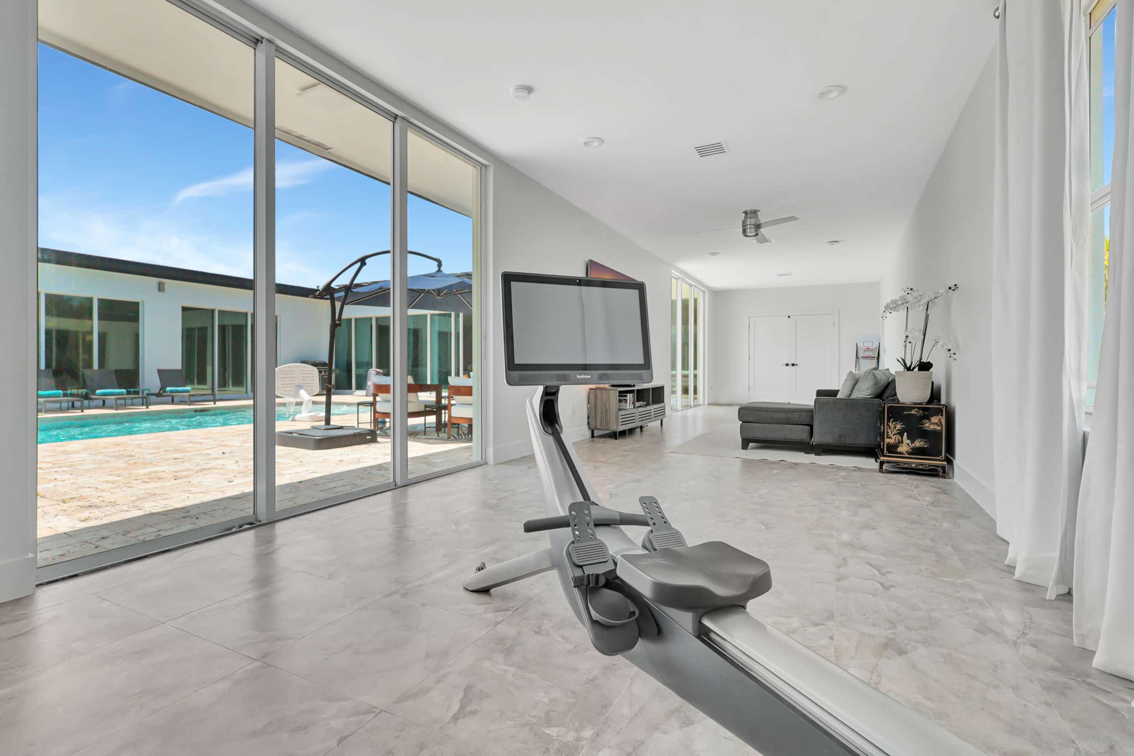 A modern indoor space featuring a rowing machine in the foreground, with large glass doors leading to a pool area outside.