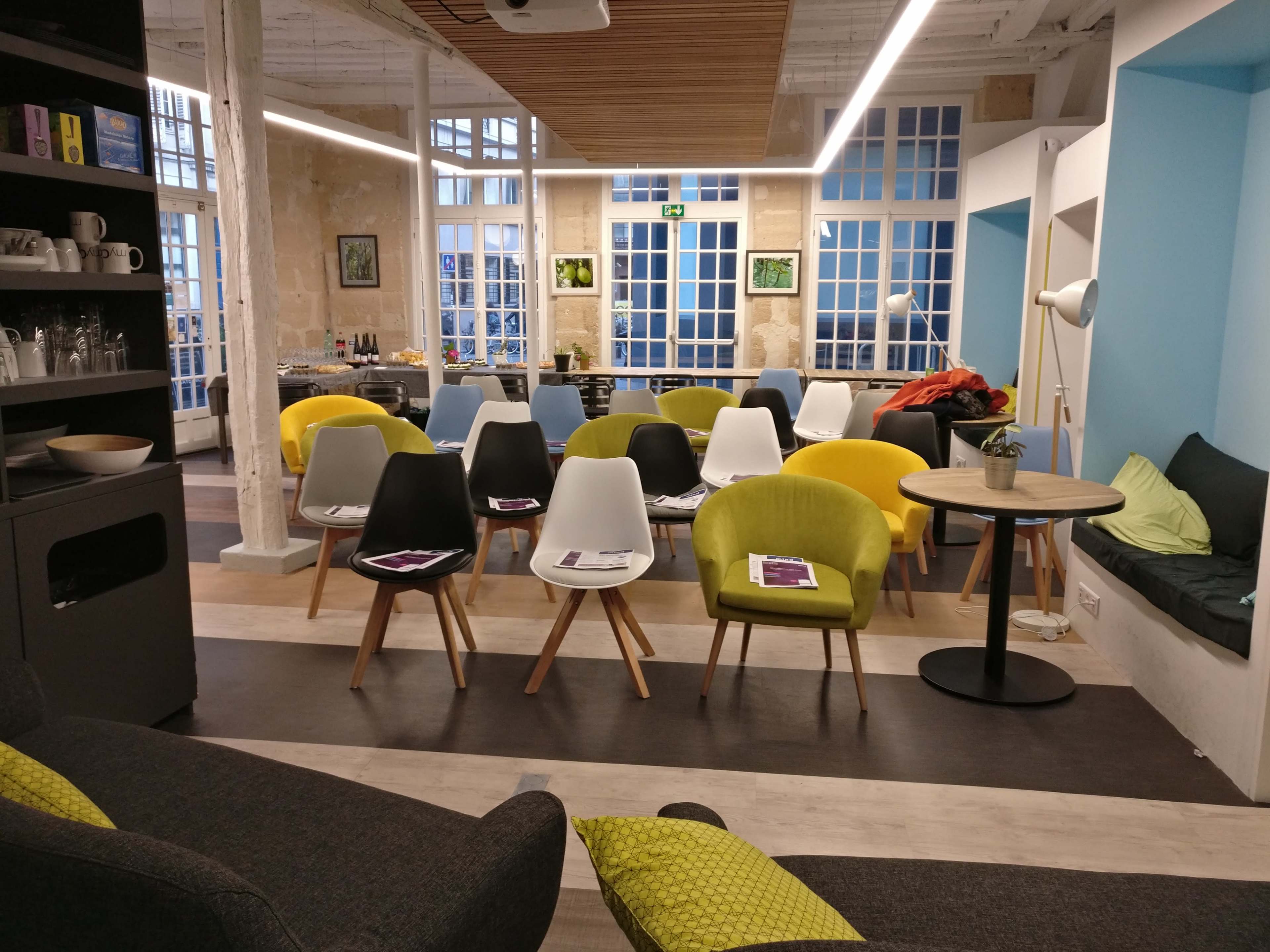 A bright, modern workspace with various colored chairs arranged around small tables, accompanied by large windows allowing natural light in.
