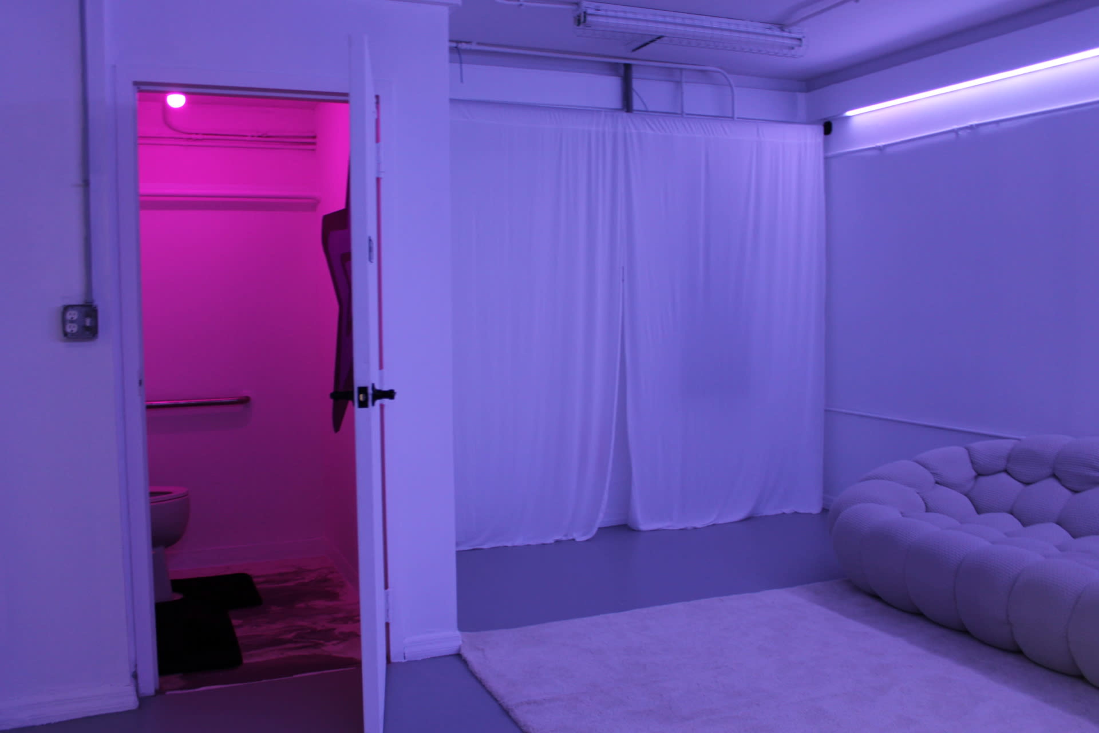 A brightly lit room features a white couch, a soft rug, and a doorway leading to a small bathroom with pink lighting.