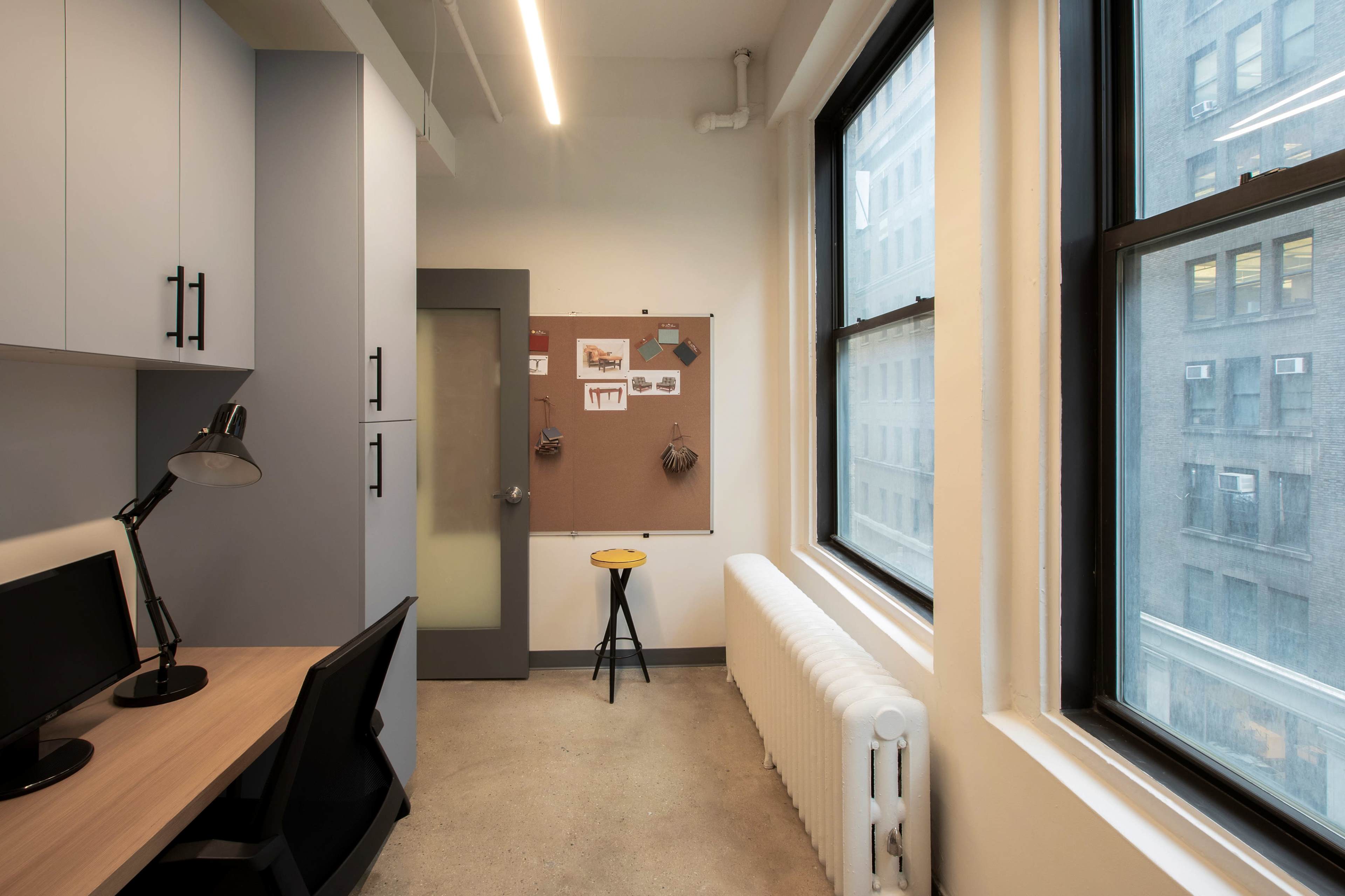 A small office space with a desk and computer on one side, a cork board on the wall with notes, and a window that views an adjacent building.