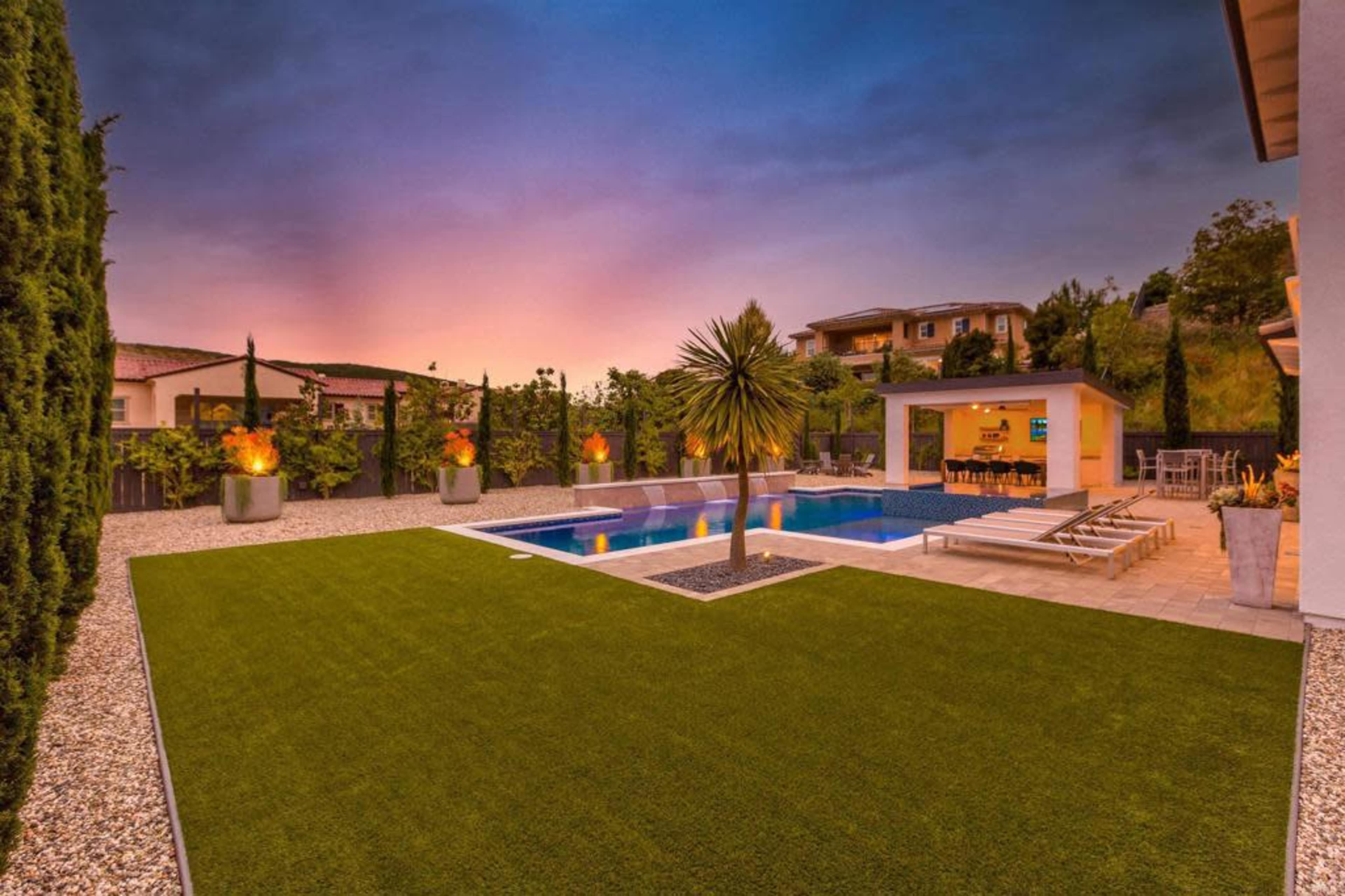 The image shows a landscaped backyard featuring a swimming pool, lounge chairs, and a dining area under an outdoor structure, set against a sunset sky.