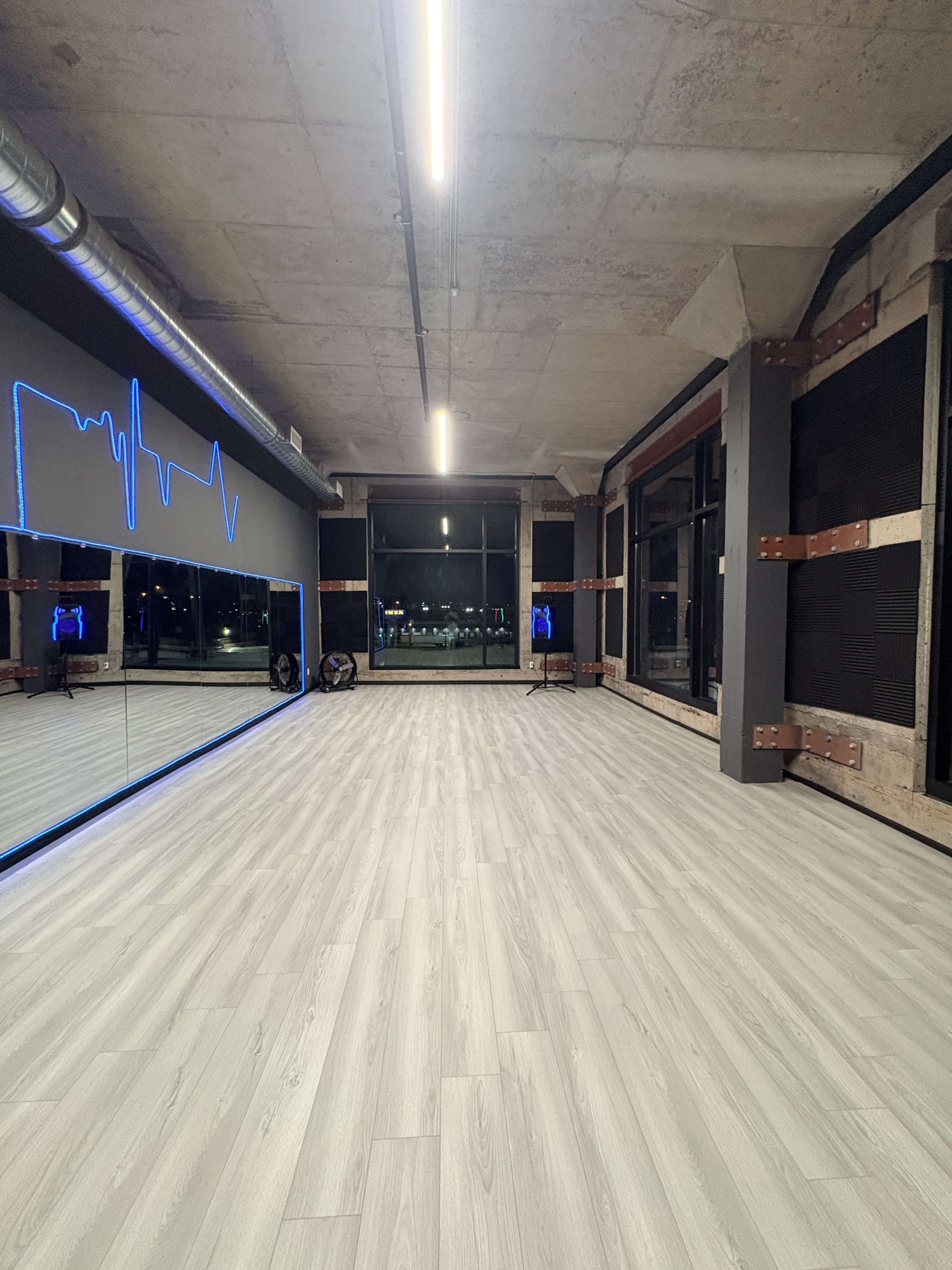 The image shows a modern dance studio with large mirrors on one wall and blue LED lighting, featuring a spacious wooden floor and industrial-style fixtures.