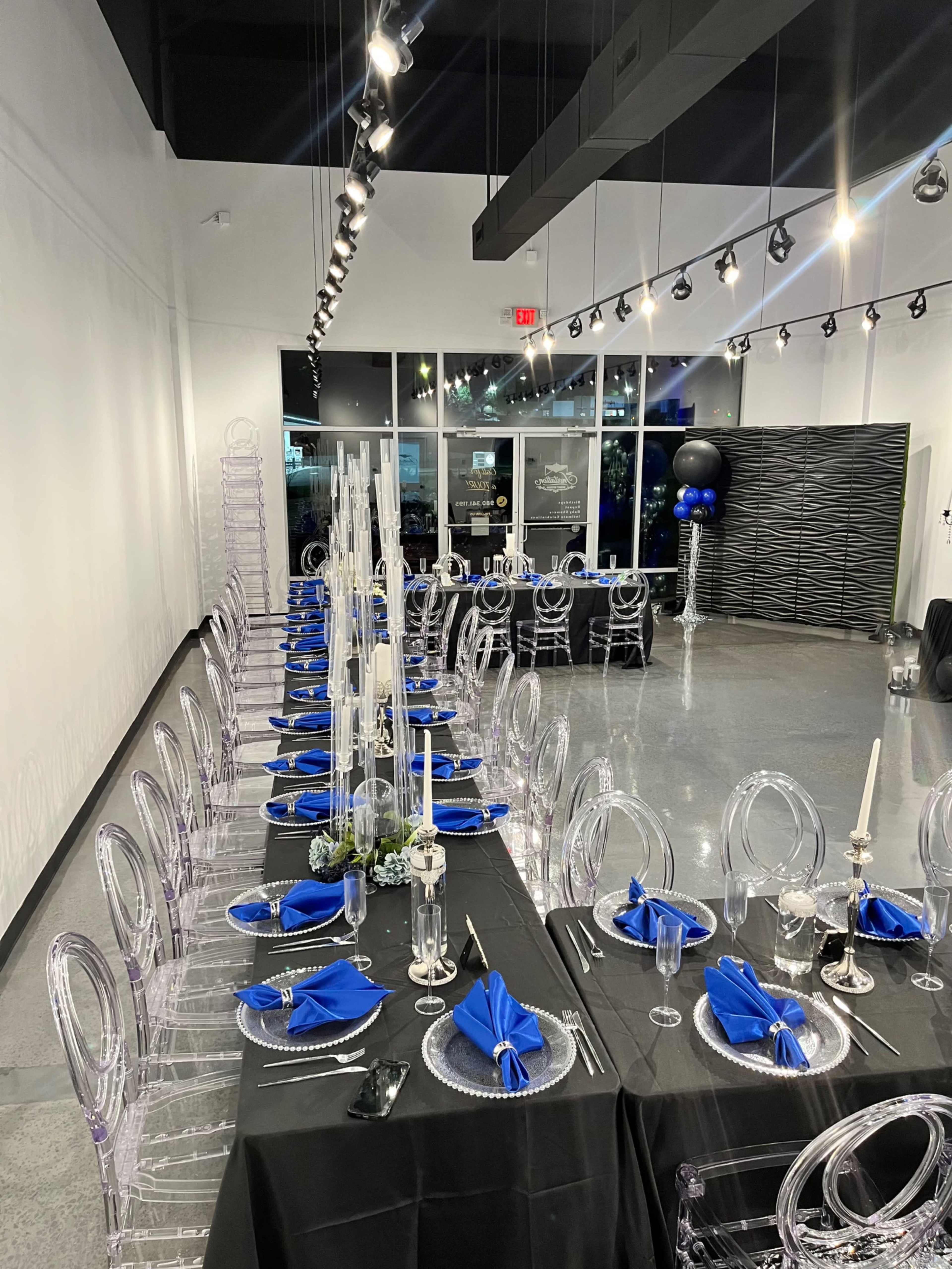 The image shows a formal dining setup with two long tables, one dressed in black linens with blue napkins and the other in clear chairs, illuminated by overhead lighting.