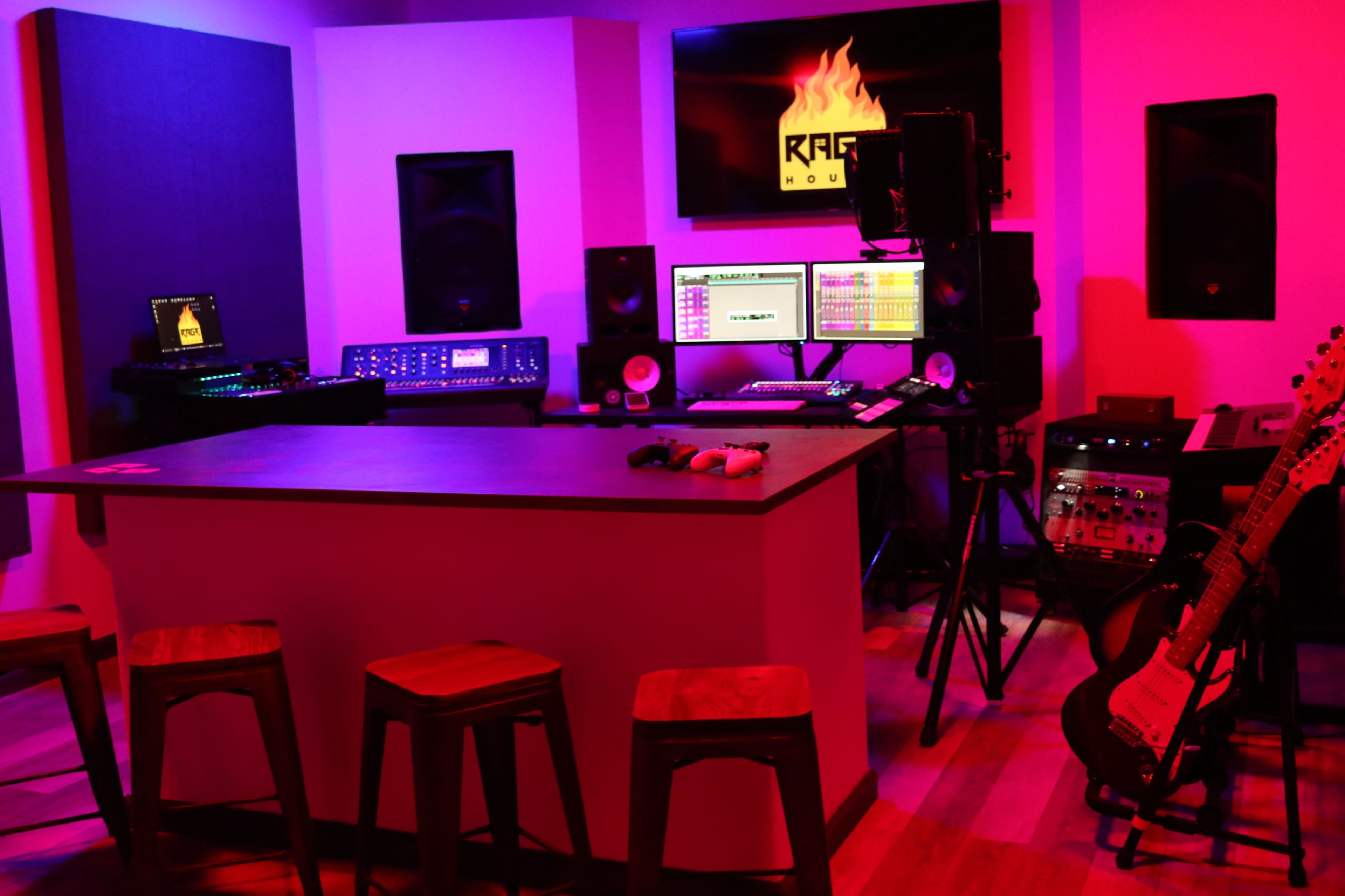 A recording studio features a workstation with computers, audio equipment, and a guitar, surrounded by seating and illuminated by colored lights.
