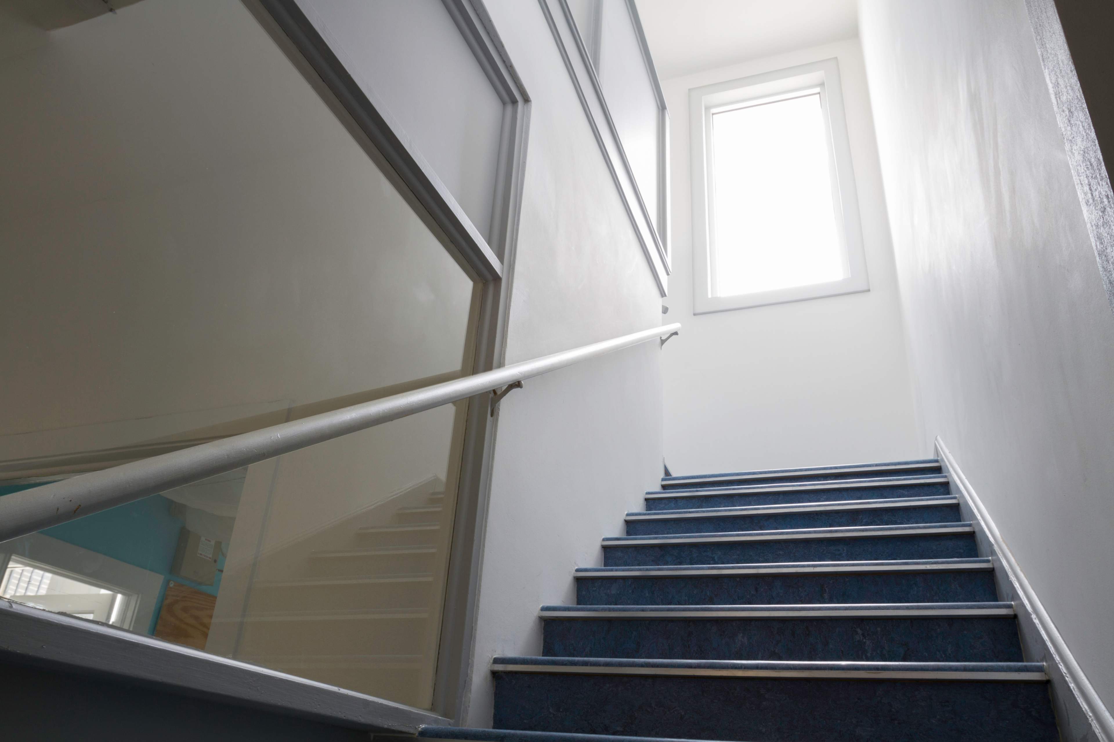 The image shows a staircase with blue steps leading up to a window at the top of a well-lit corridor.