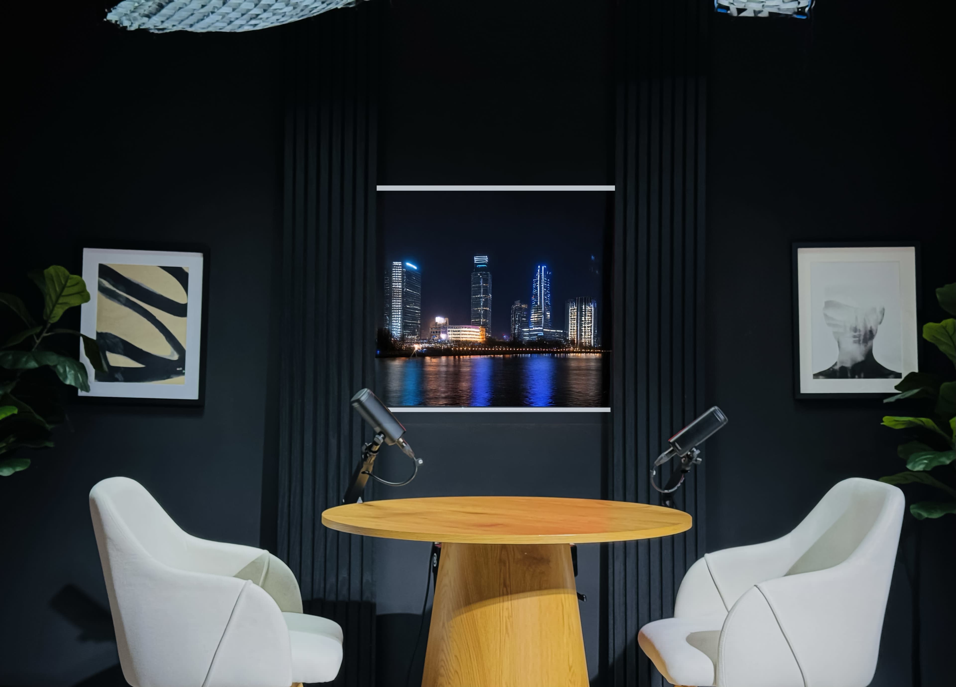 The image shows a modern studio setup with two white chairs and a round wooden table, featuring a backdrop of a city skyline at night.