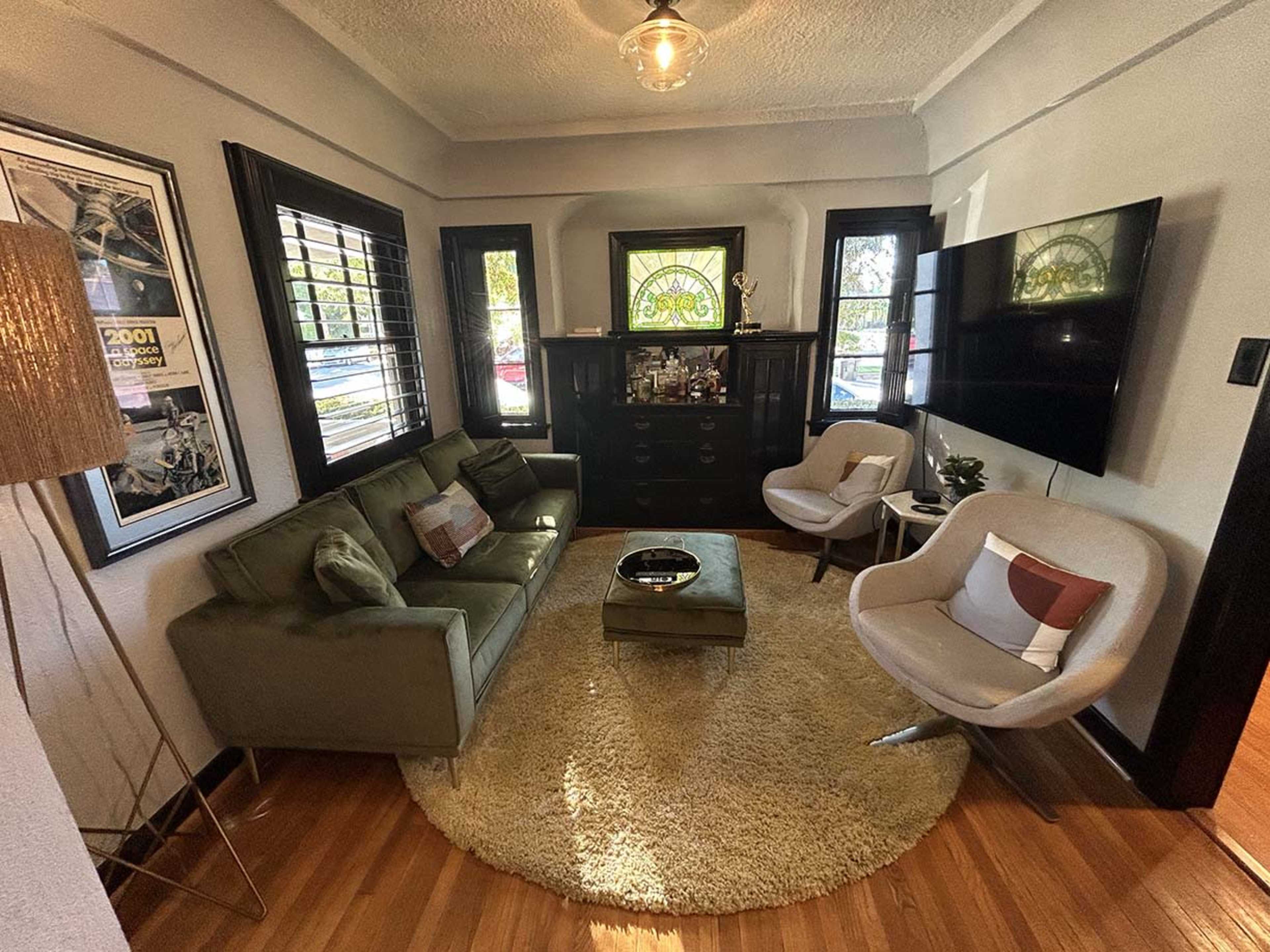 The image shows a cozy living room with a green sofa, two armchairs, a round coffee table on a circular rug, and a mounted television on the wall.