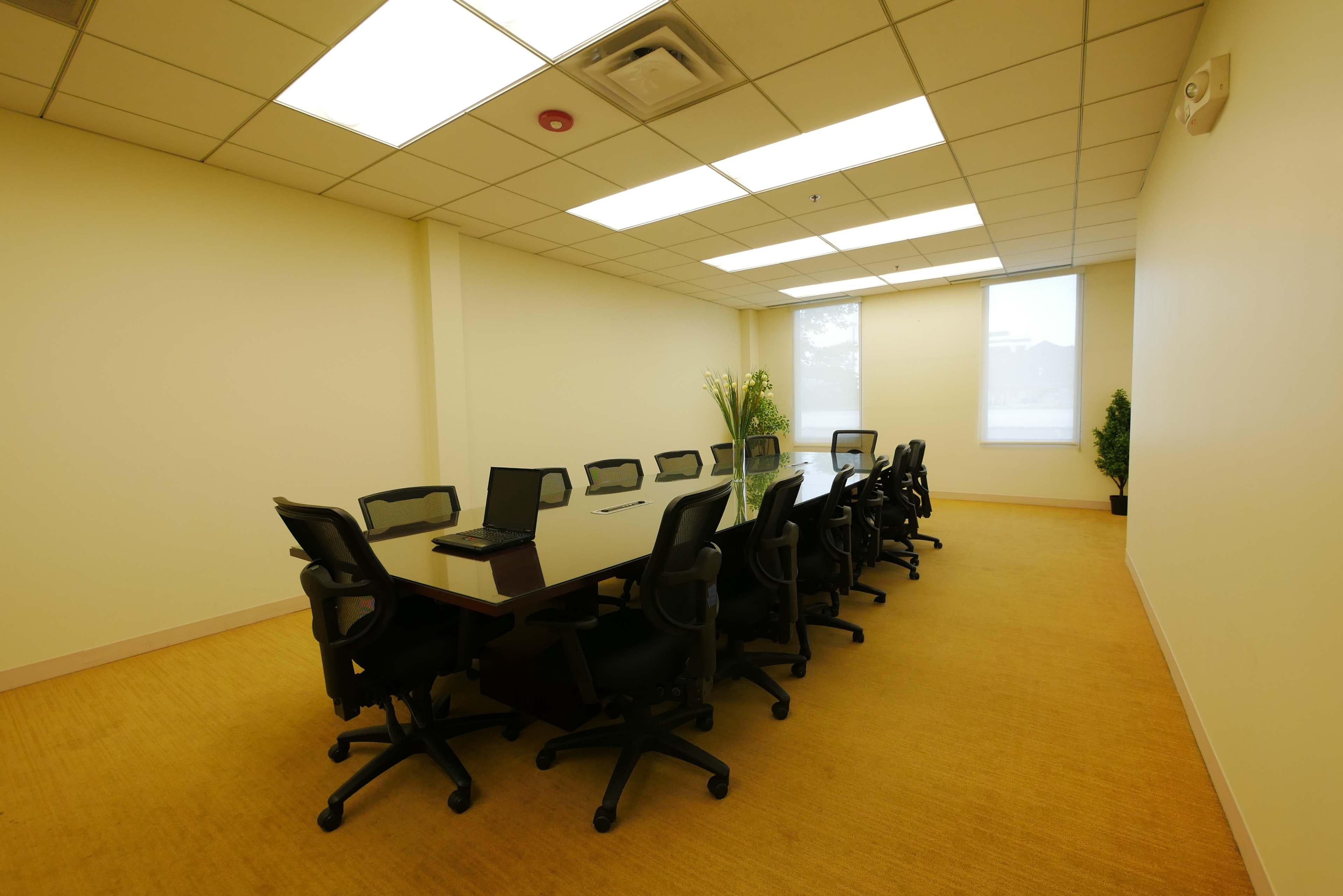 A conference room features a long table surrounded by black office chairs, with a laptop placed on one side and large windows allowing natural light.