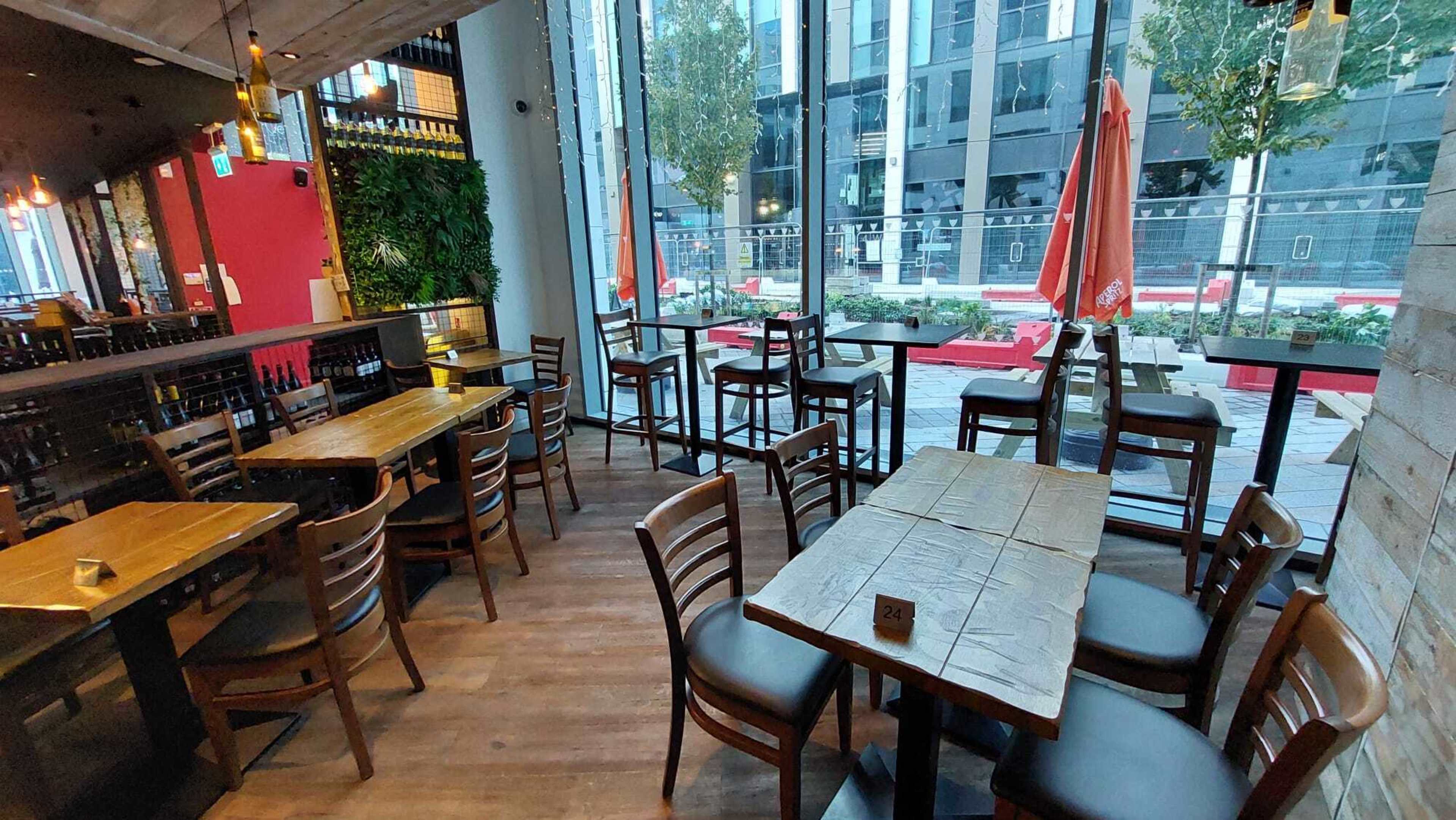 A modern restaurant interior with wooden tables and chairs arranged near large windows overlooking a city street.