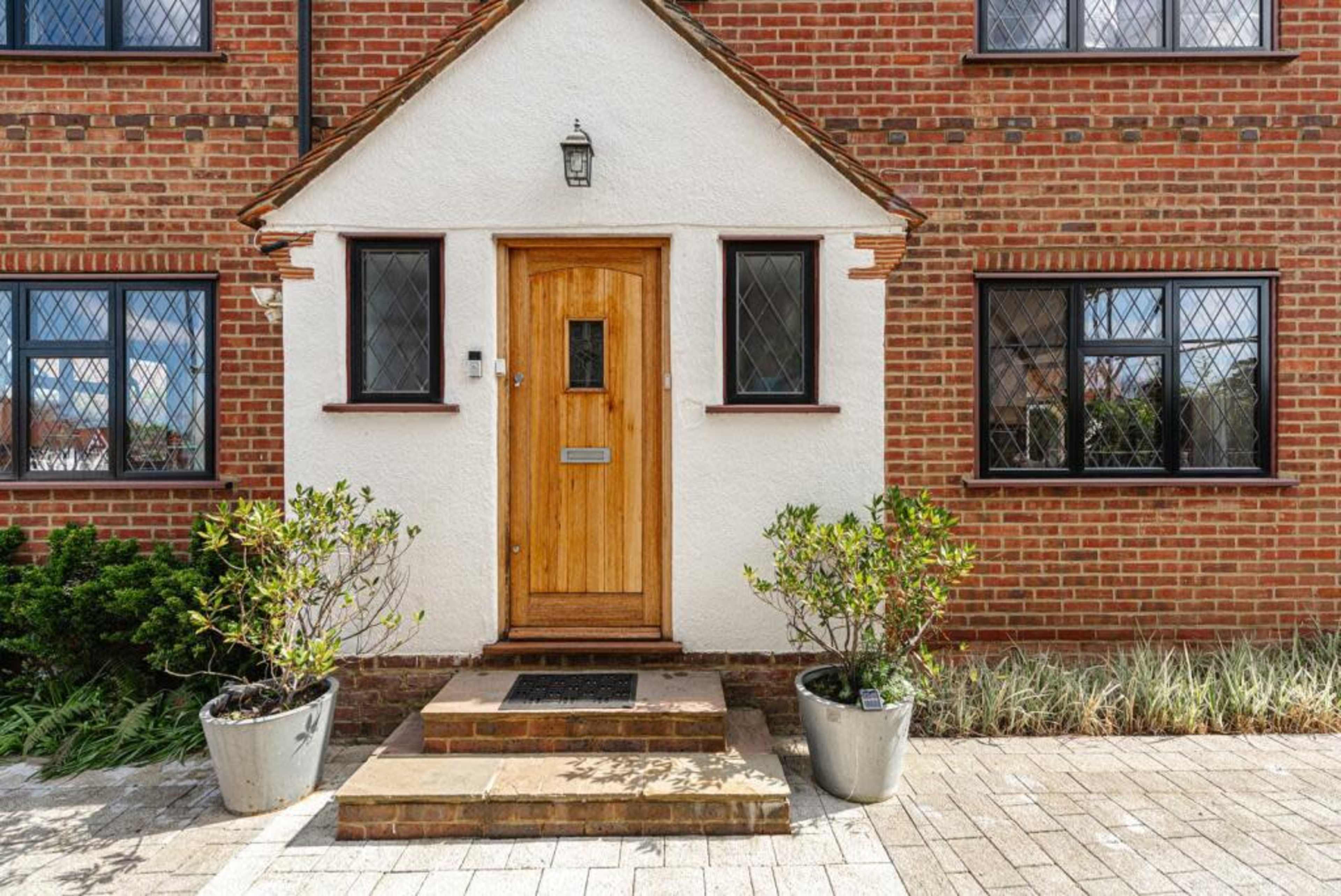 The image shows a front entrance of a brick house with a wooden door and two potted plants on either side.