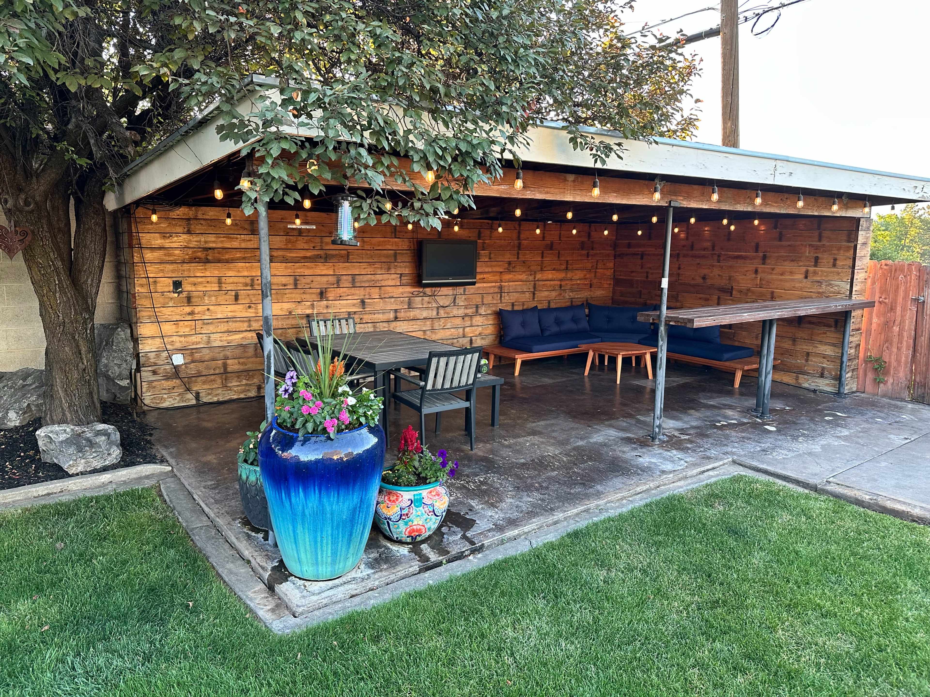 The image shows an outdoor patio area with a wooden structure, a table and chairs, a blue couch, decorative planters, and string lights overhead.