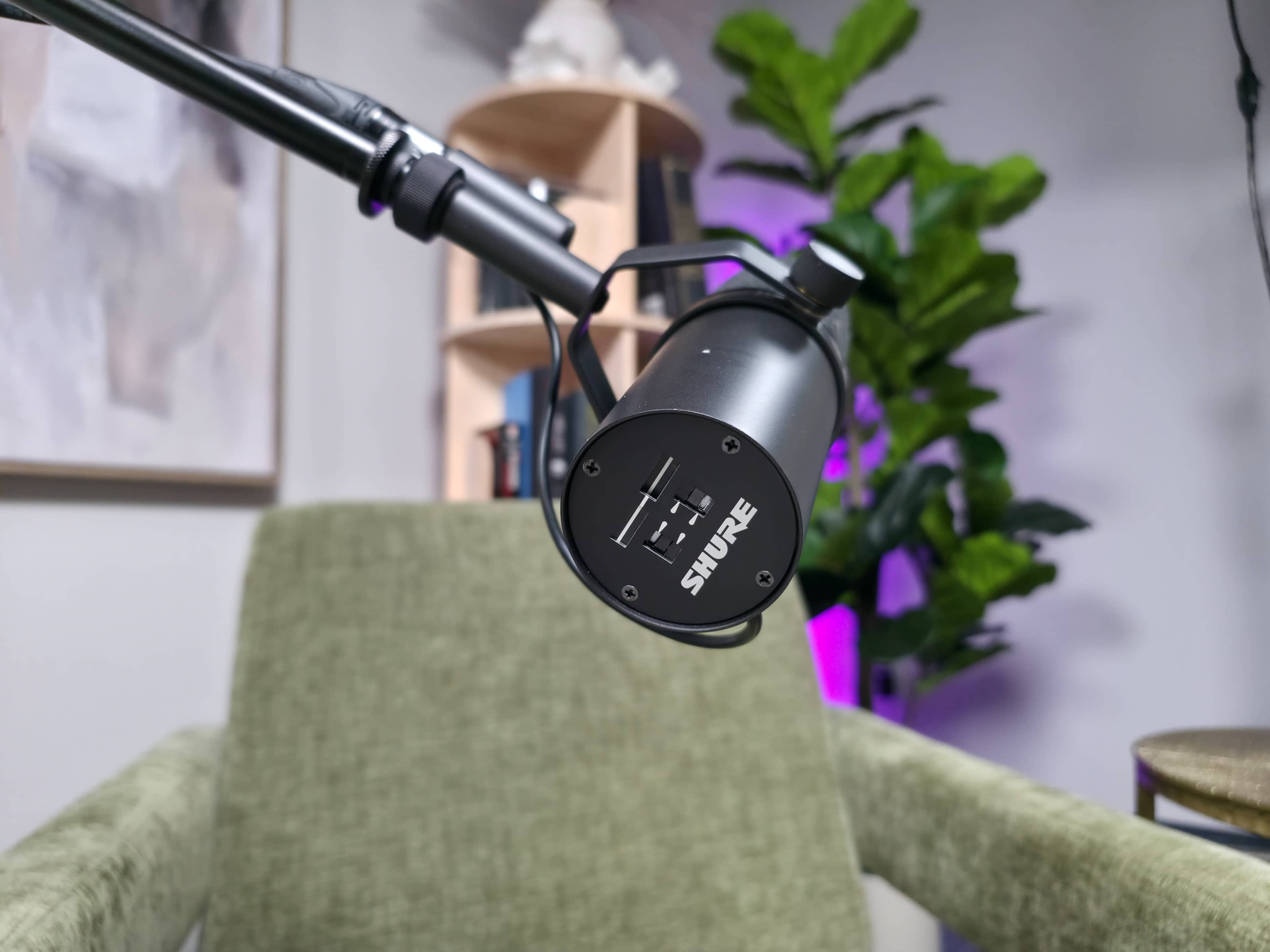 A close-up of a microphone mounted on a boom arm positioned near a green chair in a well-lit room with shelves and plants.