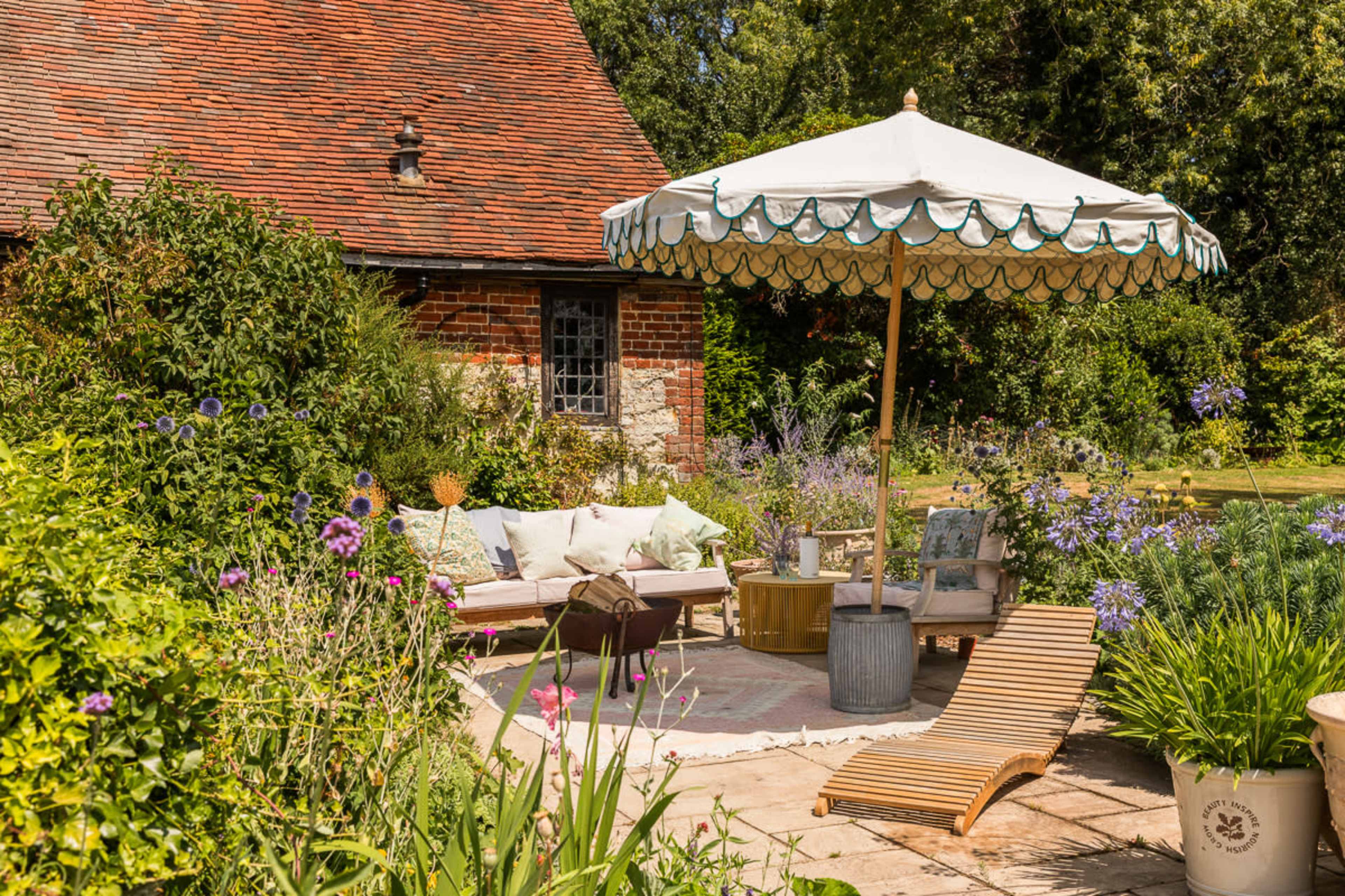 A shaded outdoor seating area with a large umbrella, surrounded by colorful flowers and greenery, features a couch, lounge chair, and a small table.