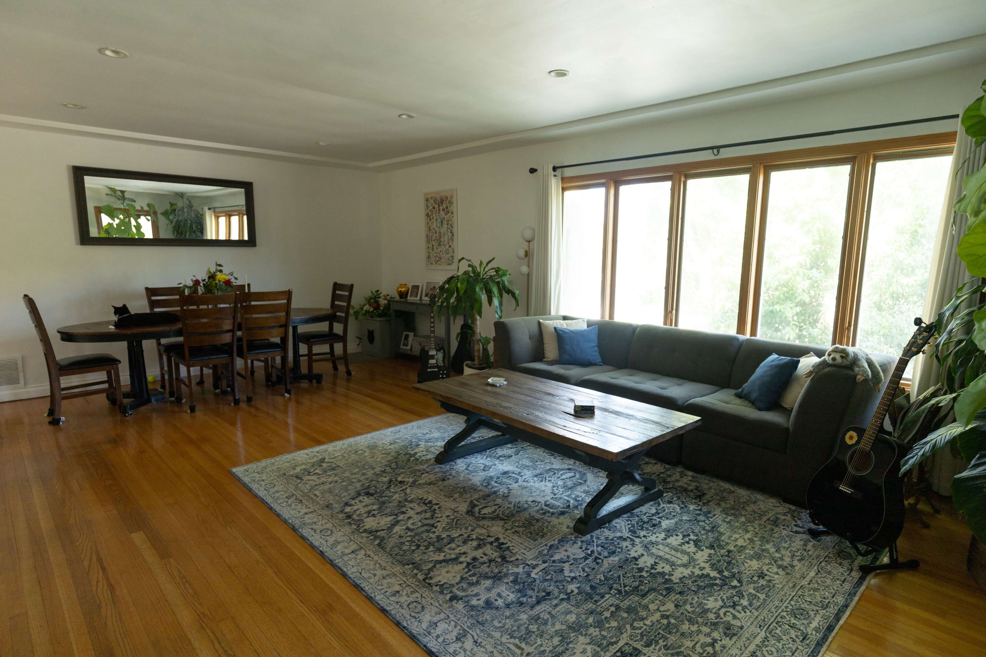 A living room features a gray sofa, a wooden coffee table, a dining area with a table and chairs, and large windows allowing natural light to enter.