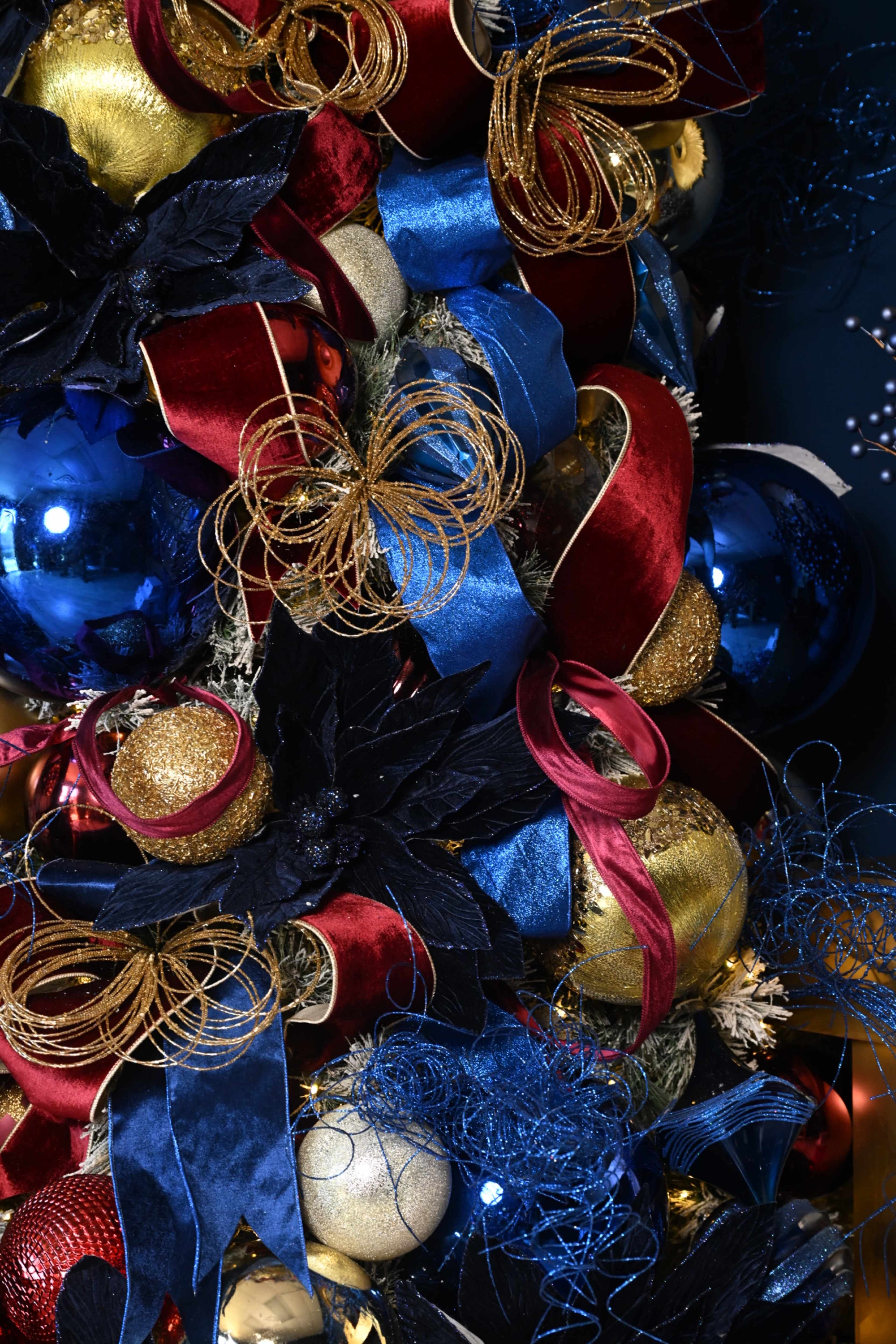 The image features a close-up of a decorated Christmas tree adorned with shiny blue and gold ornaments, red and blue ribbons, and dark foliage accents.