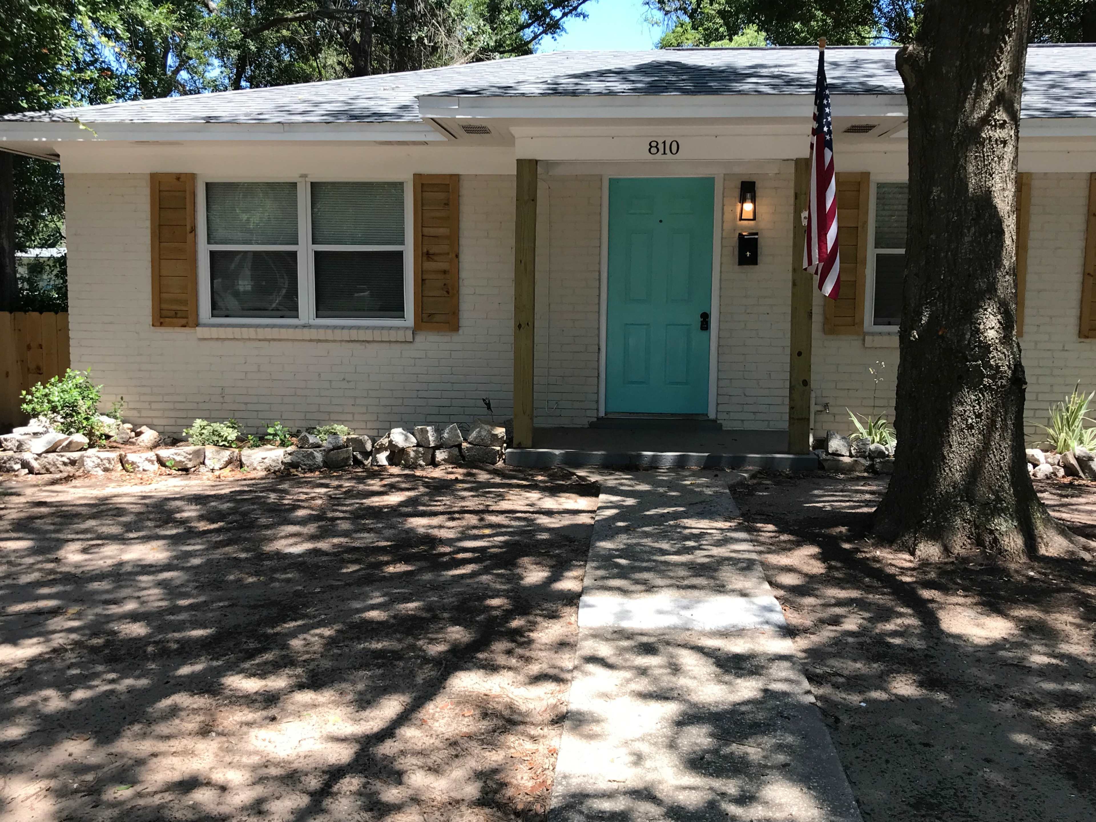 A single-story white brick house features a turquoise front door, with wooden shutters and an American flag hanging beside it.