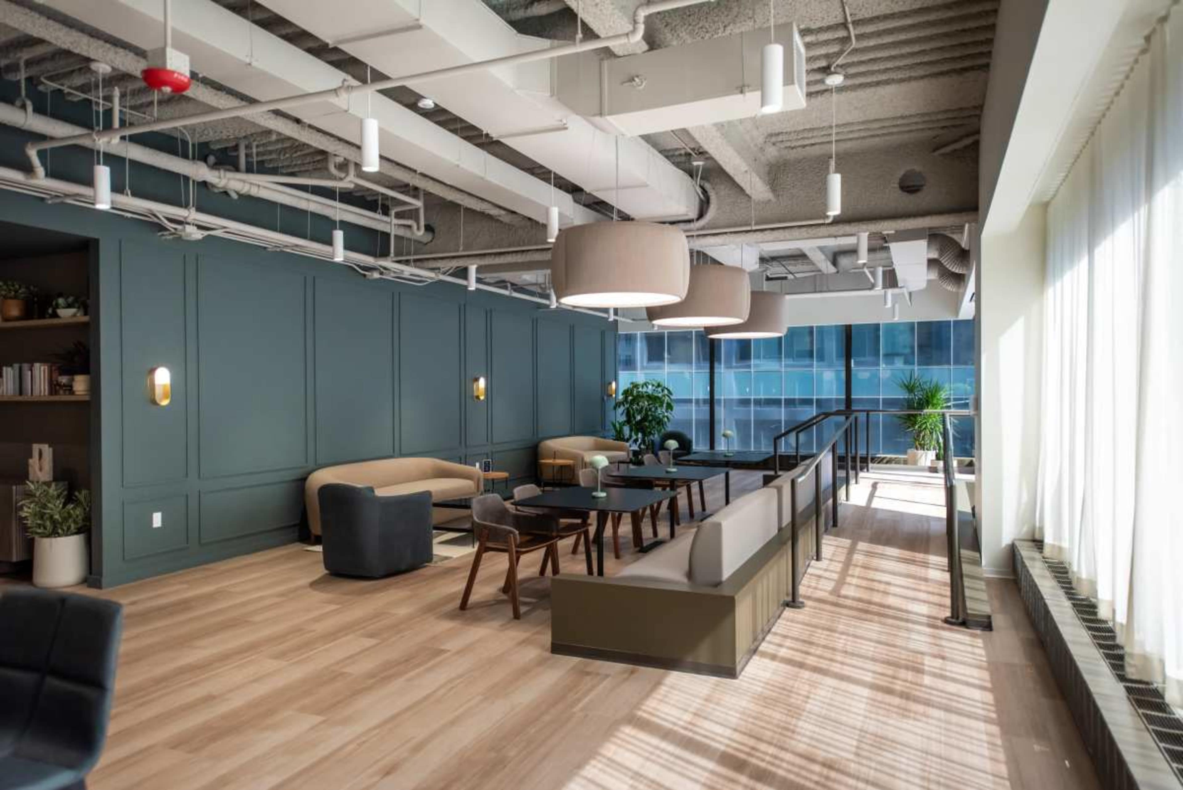 The image shows a modern lounge area with large windows, featuring comfortable seating, a coffee table, and greenery throughout.