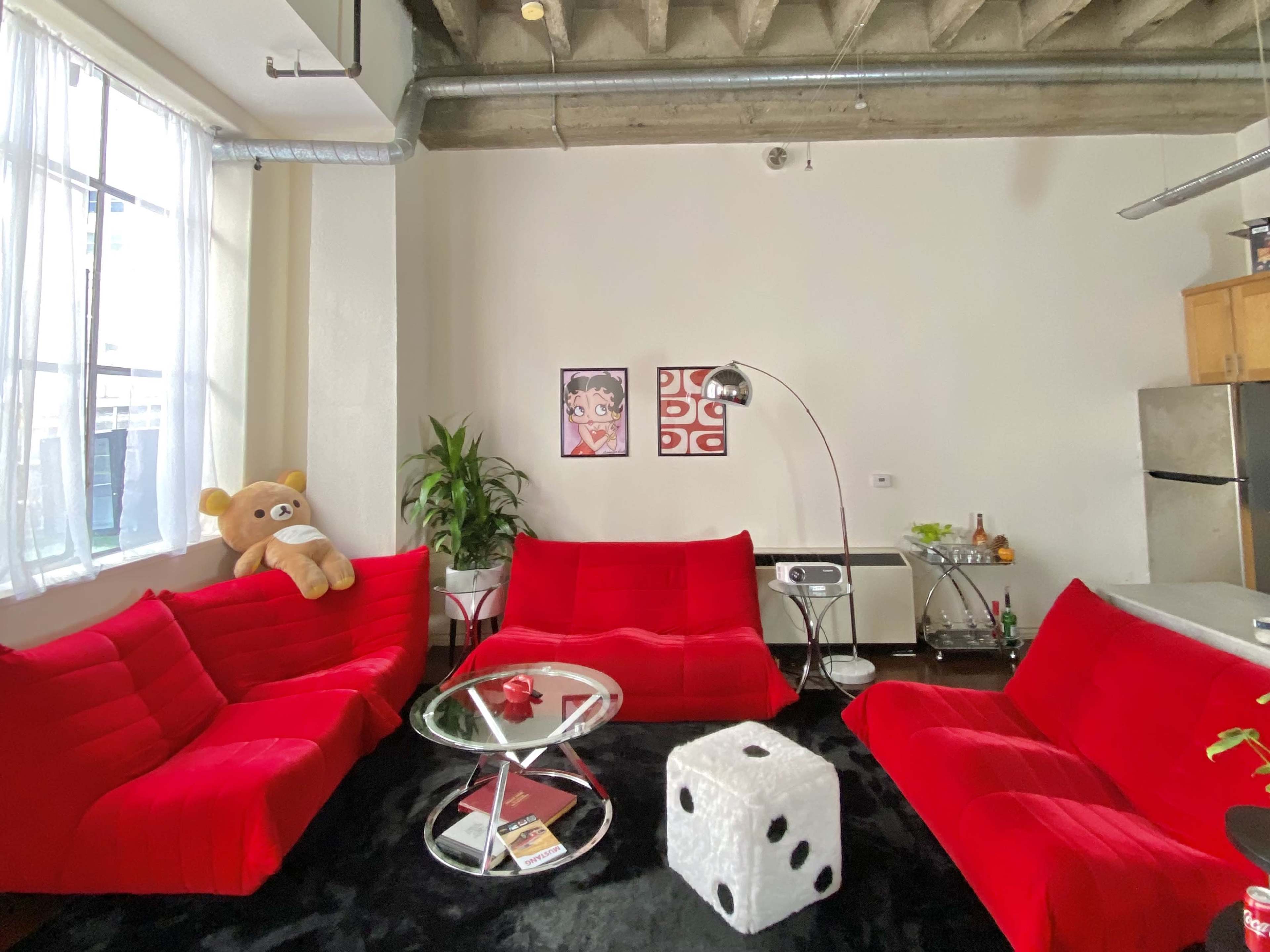 The image depicts a modern living room featuring red sofas, a glass coffee table, a plush bear, and artwork on the walls.