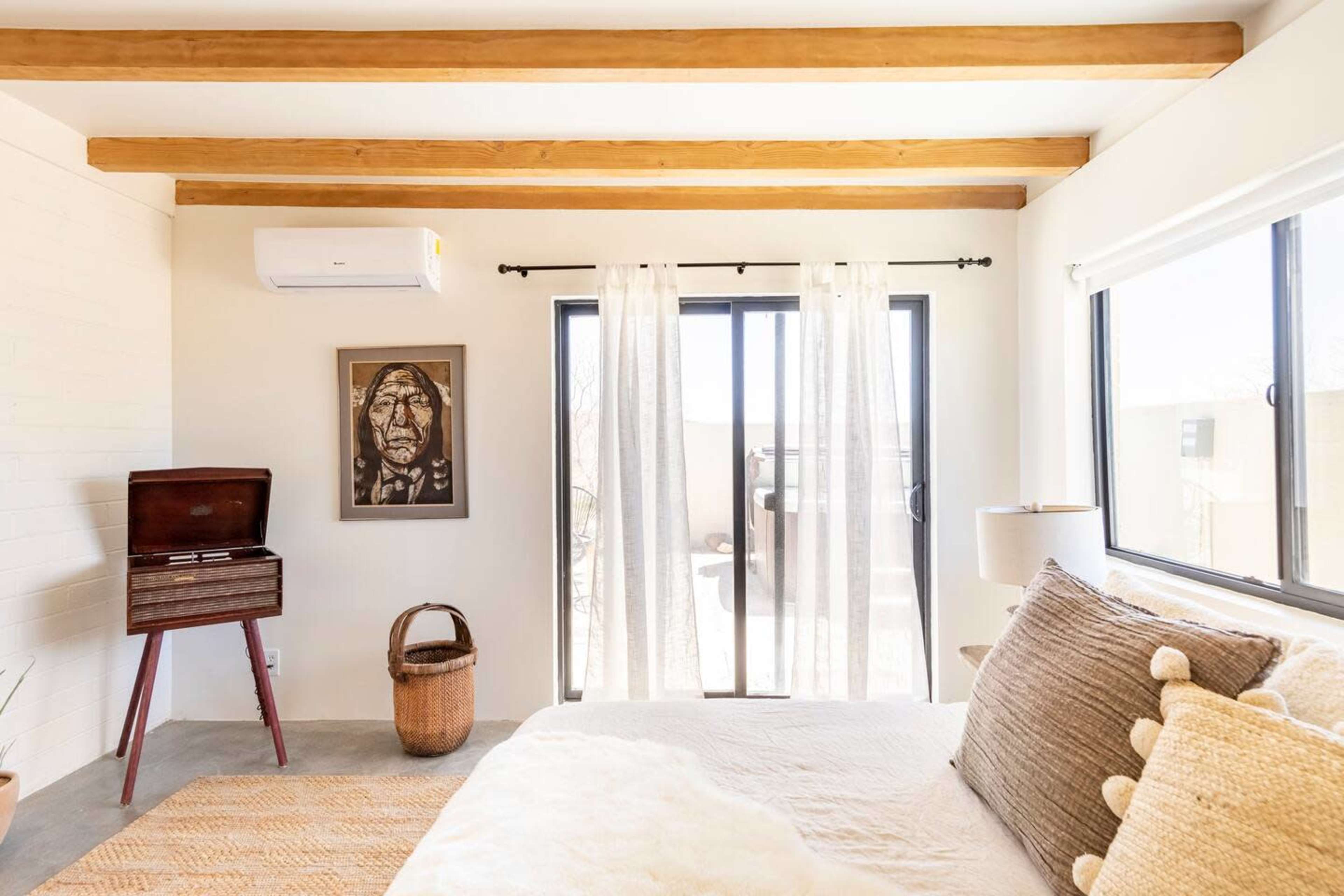 A bright bedroom featuring a bed with textured linens, a vintage record player, a woven basket, and large windows with sheer curtains.